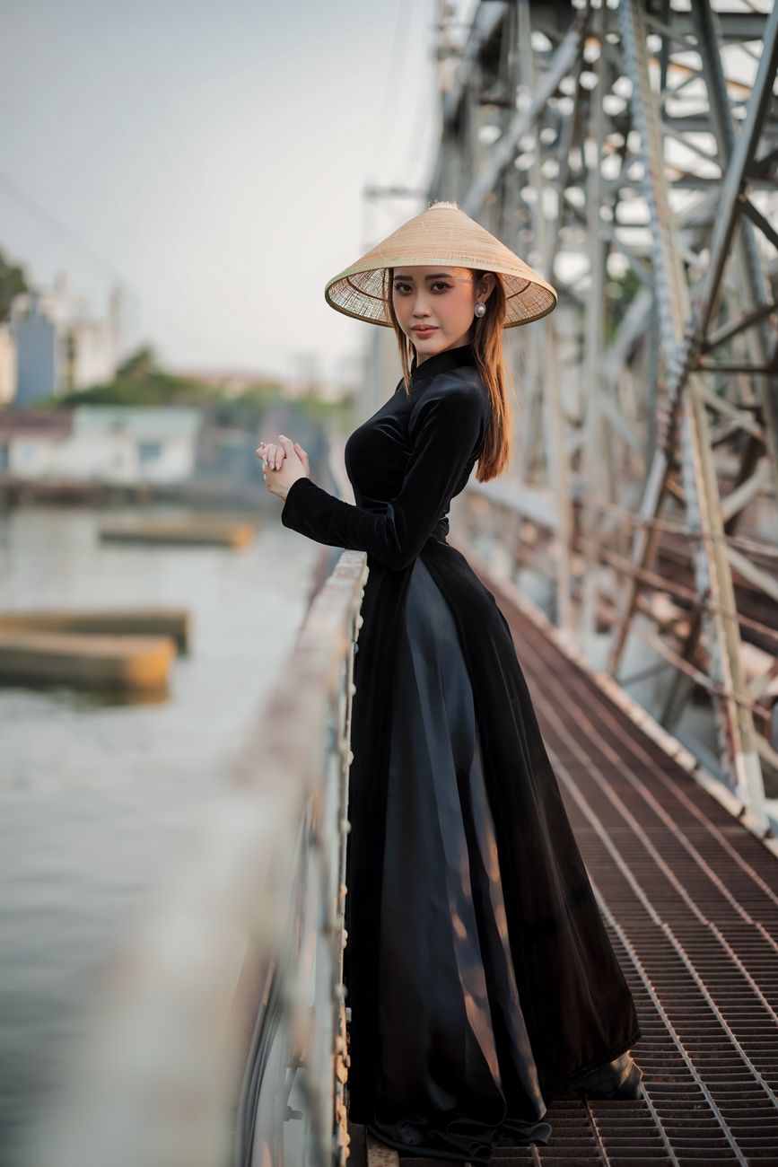 Stylish woman in Vietnamese ao dai with a conical hat standing on a steel bridge, capturing cultural elegance.