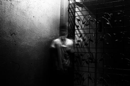 一张灰色照片中，一个模糊的人物置身于怪异的室内环境，周围散落着金属制品