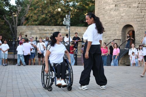 Une femme en fauteuil roulant et une autre femme debout se regardent et sourient sur une place pavée, devant un mur de pierre ancienne, entourées par plusieurs personnes dans un parc.