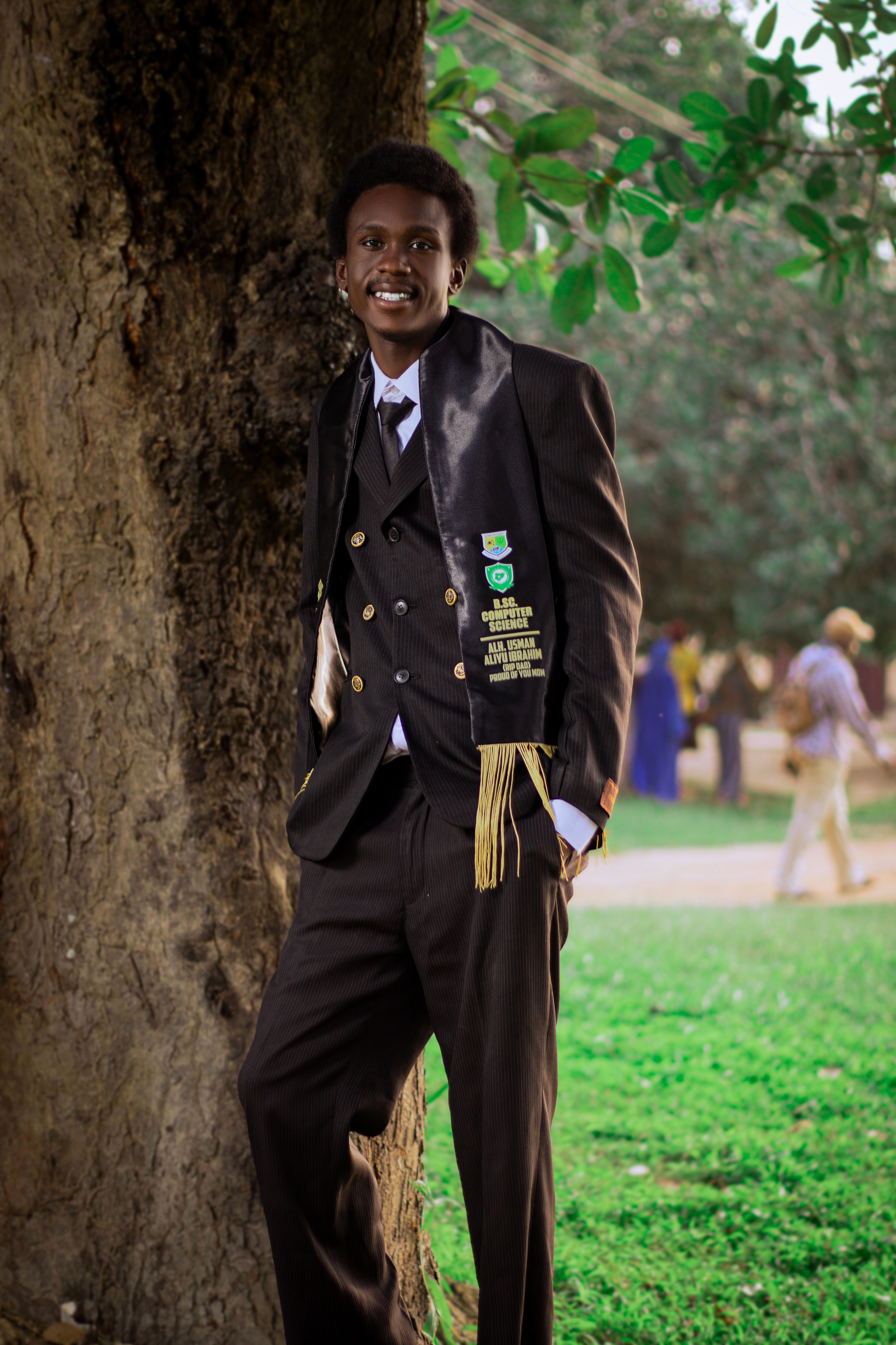 Smiling graduate poses outdoors in suit and sash, celebrating achievement.