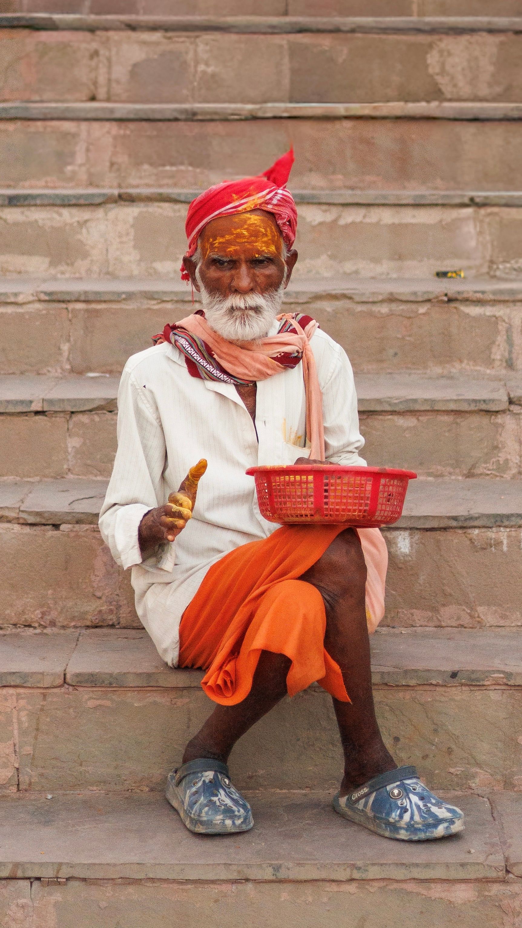 Elderly man in vibrant traditional attire sitting on stone steps in Varanasi, India, showcasing cultural richness.