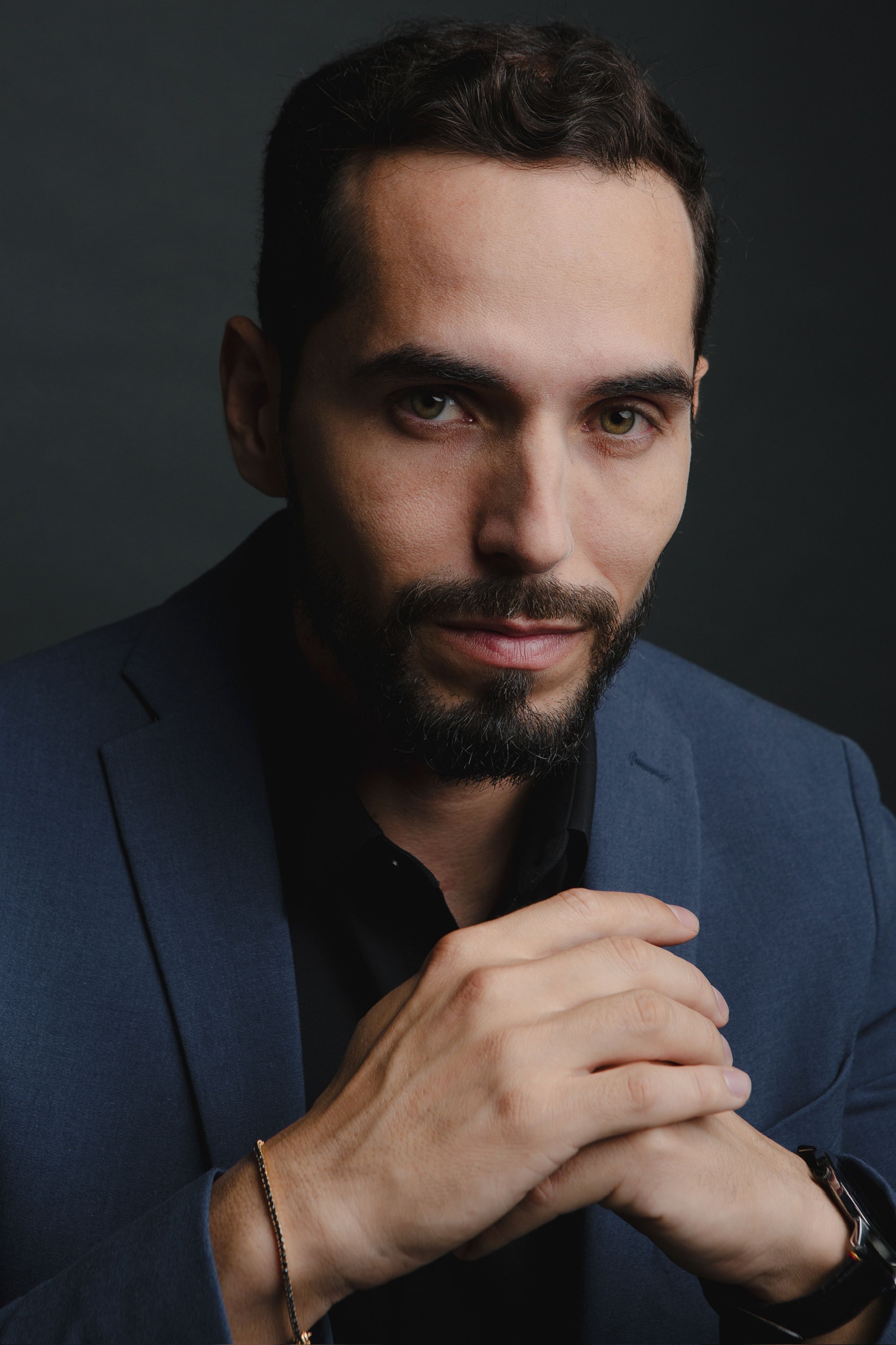Close-up portrait of a confident man with a beard, wearing a blue suit against a dark background.
