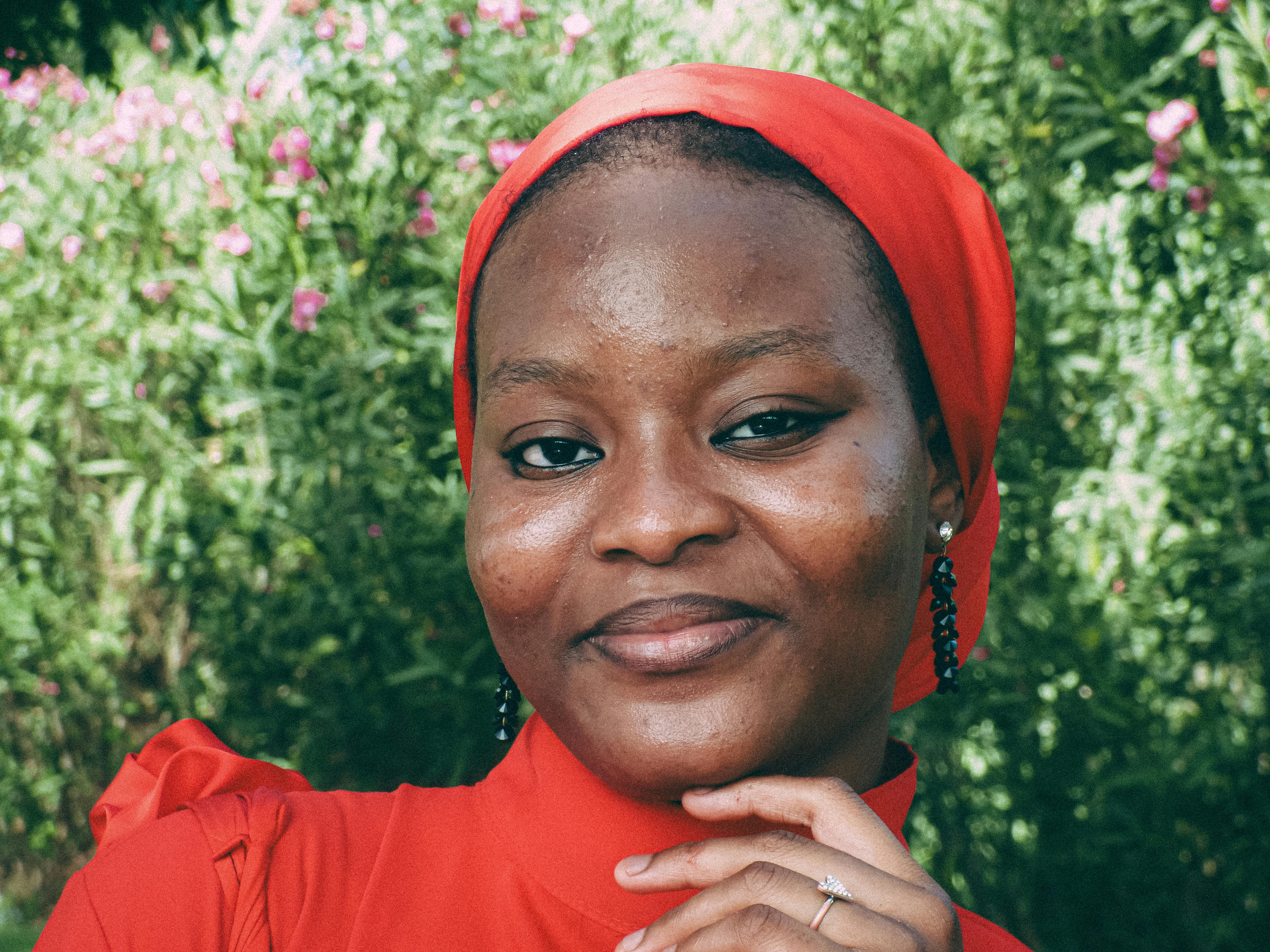 A serene woman in a red headscarf smiles against a lush green backdrop during the day.
