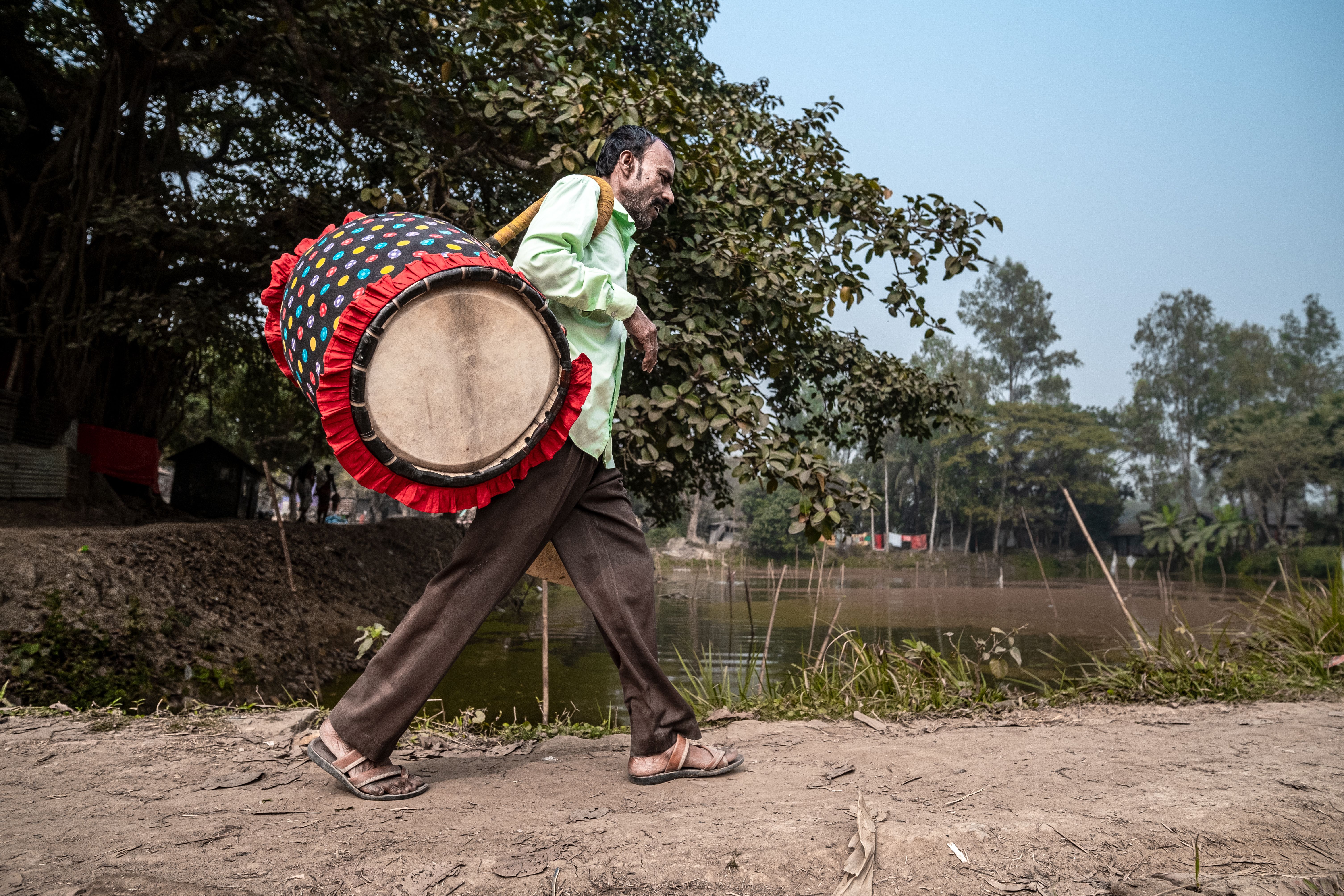 A man carrying a decorated drum walks along a rural path in a serene outdoor setting.