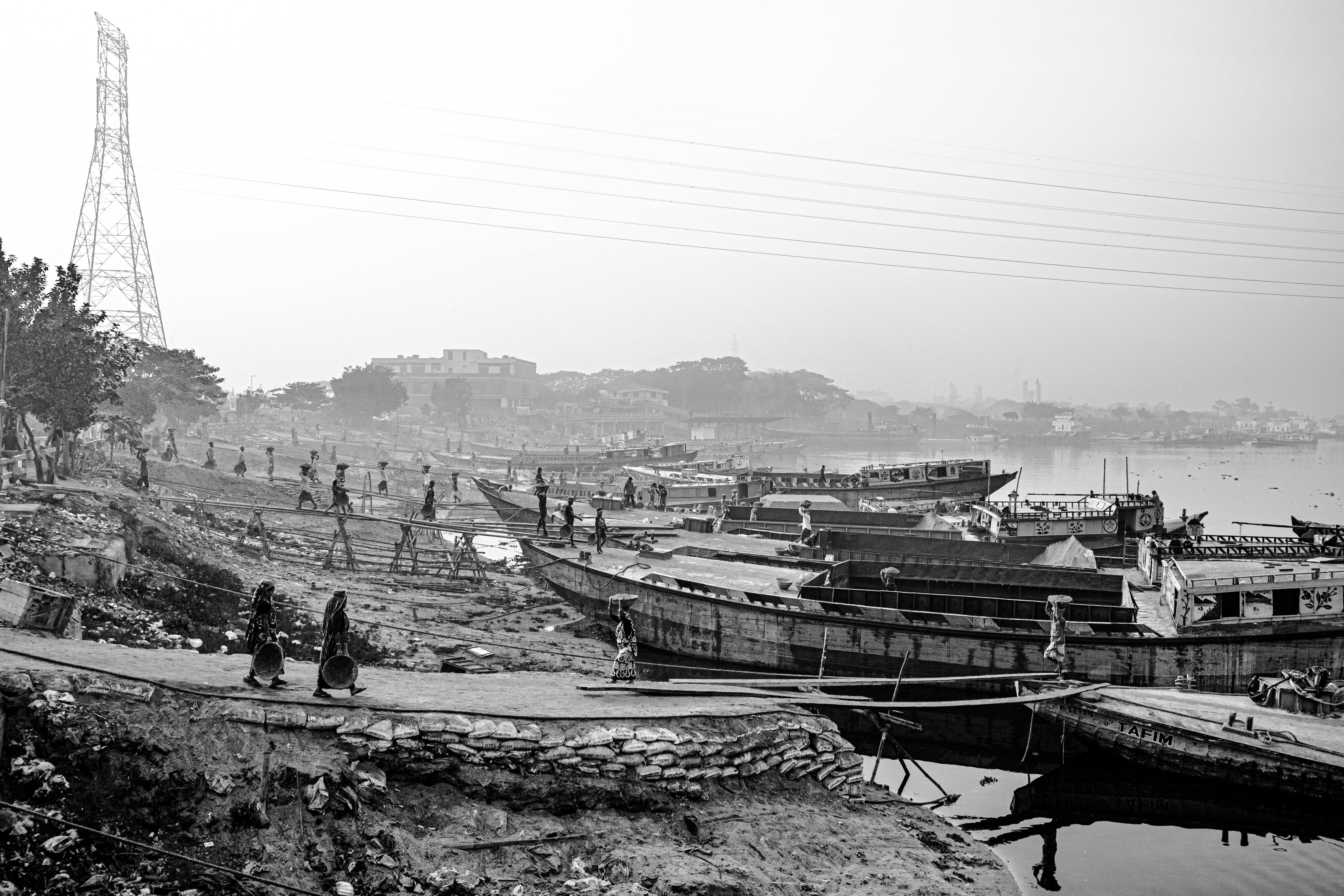 Monochrome image of a bustling riverbank with boats and people during the day.