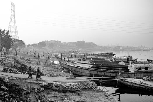 Monochrome image of a bustling riverbank with boats and people during the day.