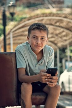 Teen boy in casual outfit sitting outside, holding a smartphone and smiling.