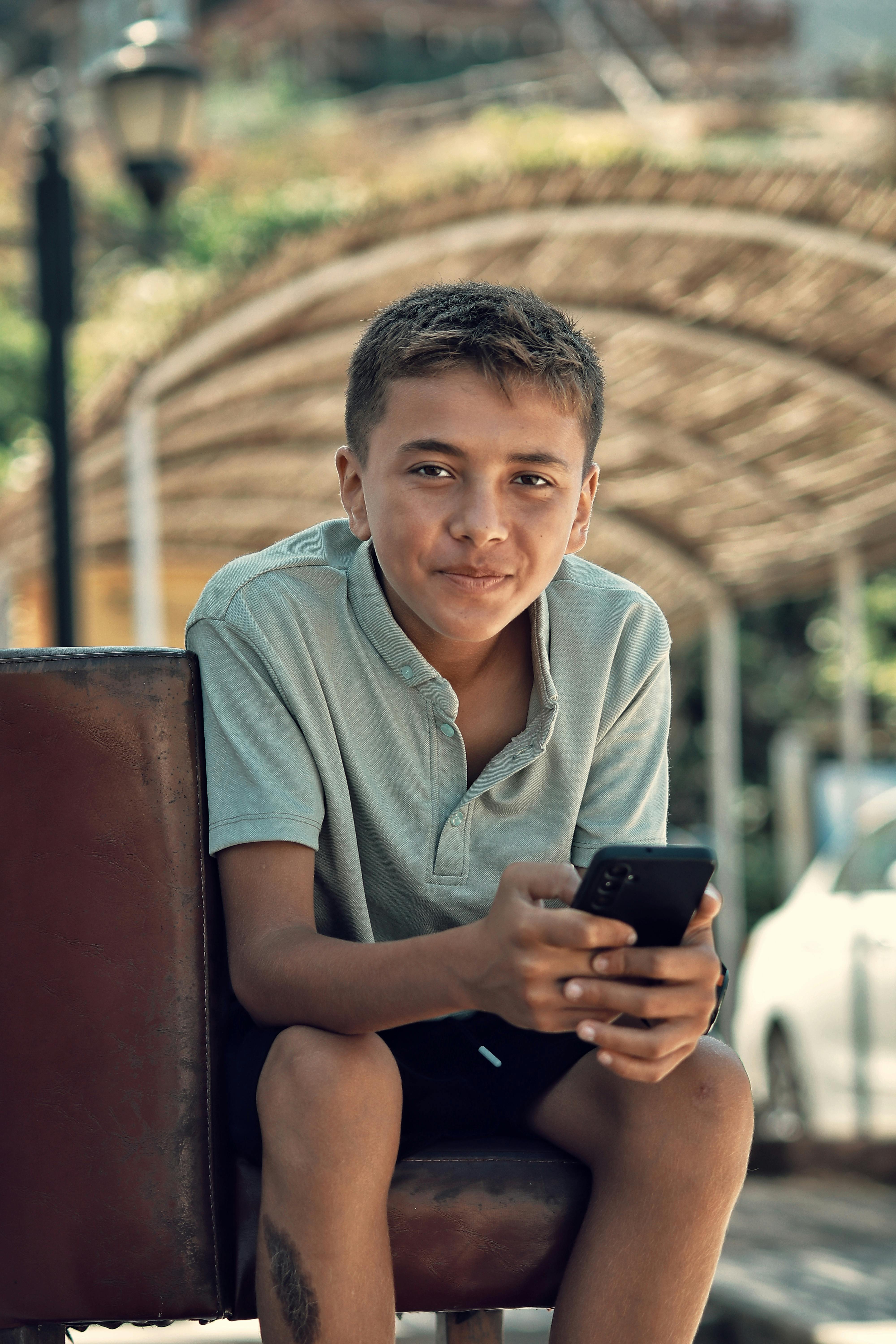 grátis Adolescente em roupa casual sentado do lado de fora, segurando um smartphone e sorrindo. Foto profissional