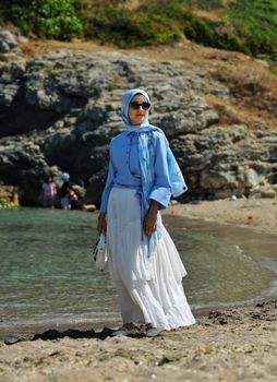 A fashionable woman in a light blue hijab and white skirt stands by a rocky beach, exuding grace and style.