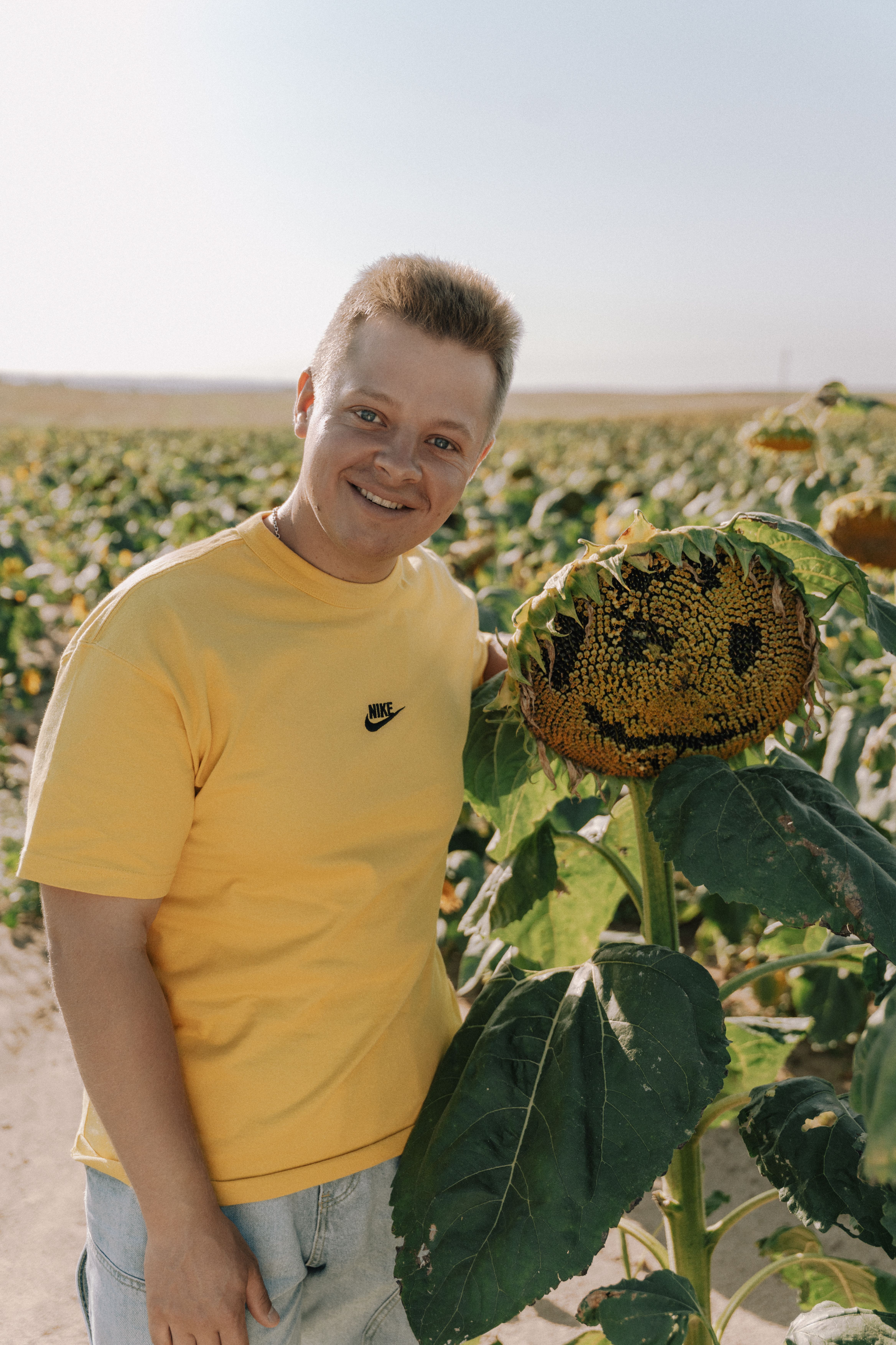 Young man in a yellow shirt smiling next to a sunflower in a sunny field.