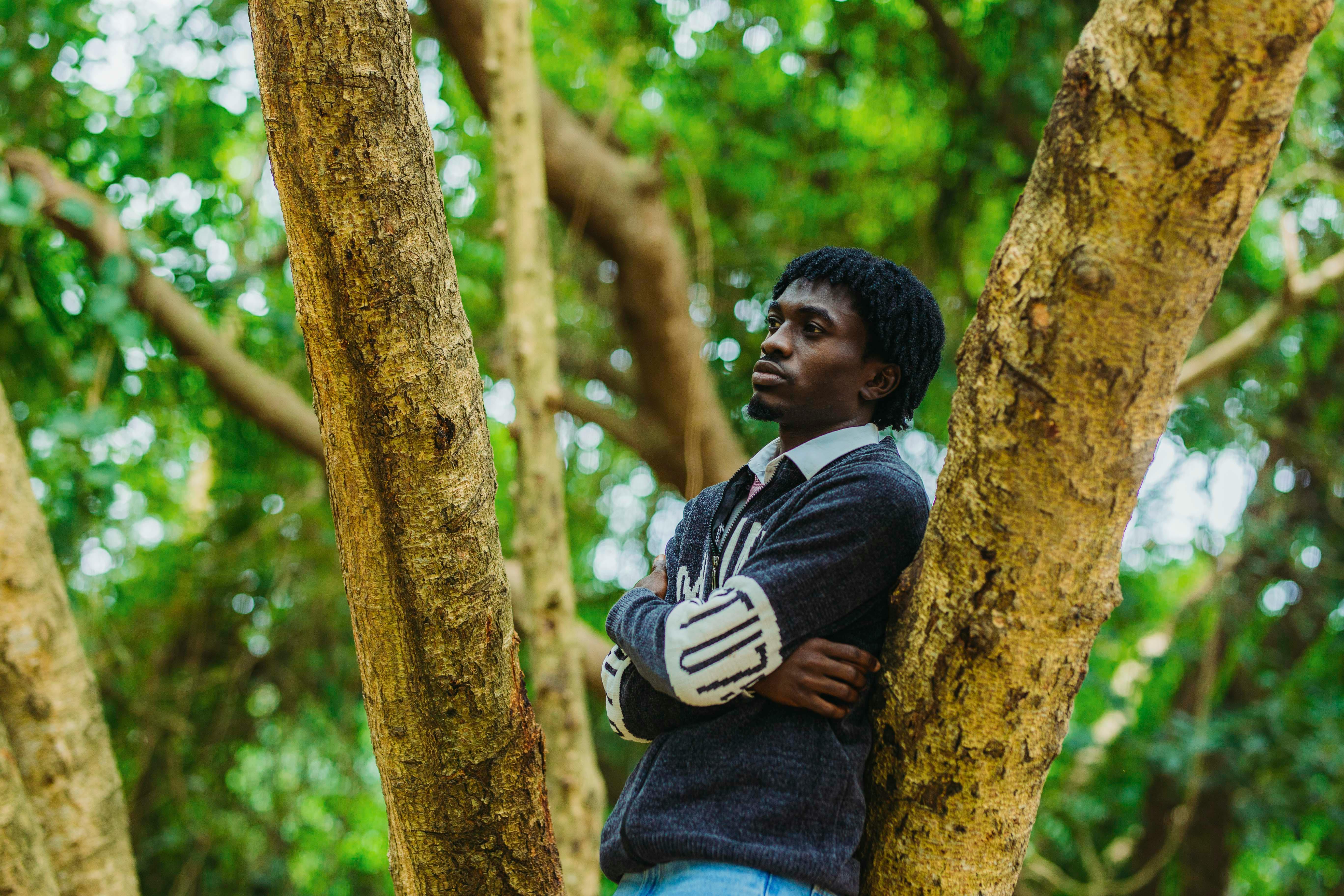 A thoughtful man leans against a tree in Accra's lush outdoor setting, blending nature and urban life.