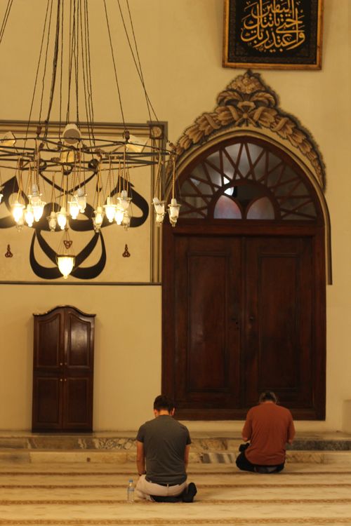 Free Two men seated in prayer inside a historic mosque, under elegant lighting and ornate designs. Stock Photo
