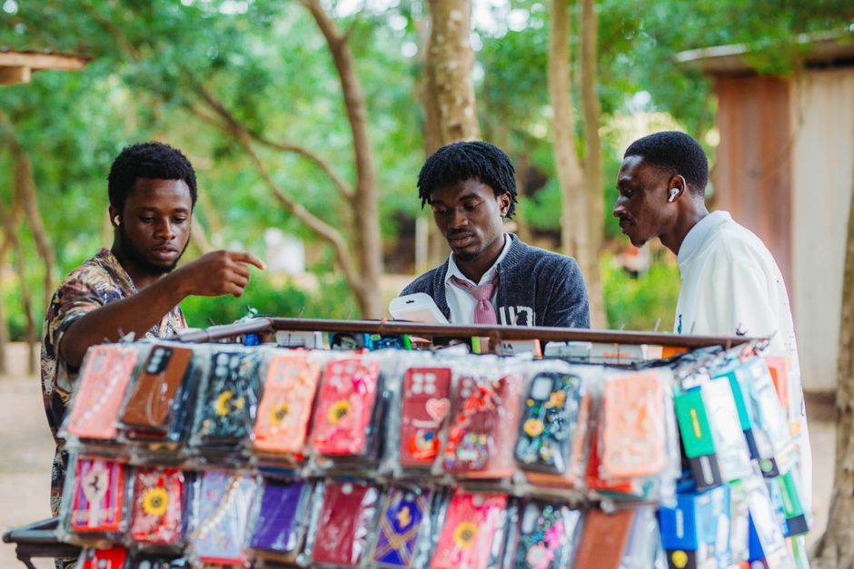 Three men browsing phone cases at an outdoor market stall in Accra, Ghana.
