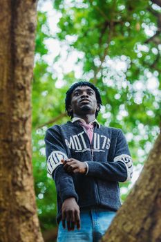 A man in casual attire stands thoughtfully between tree trunks amidst lush green foliage in Accra.
