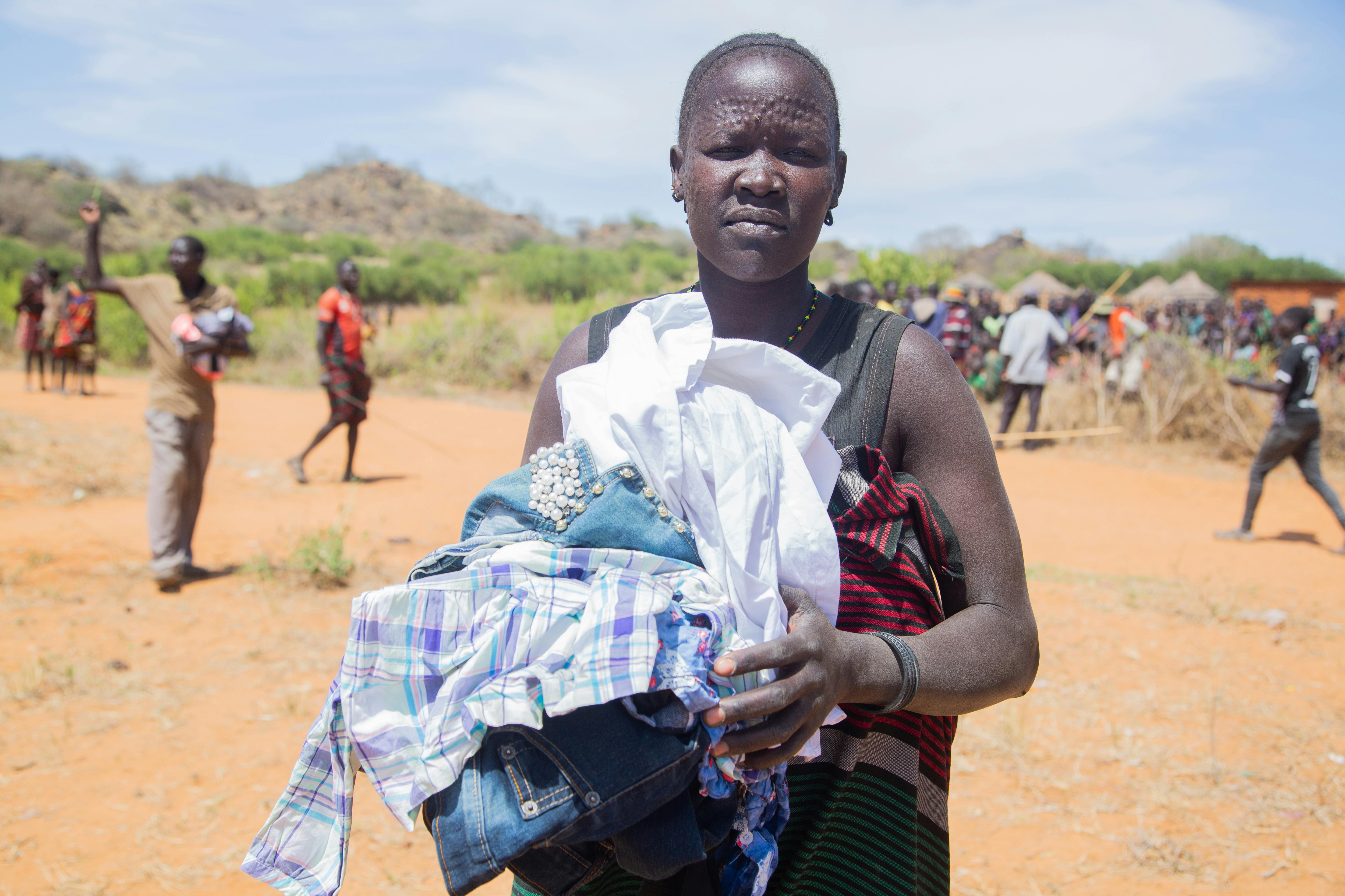 A woman in a remote village holds clothing, surrounded by villagers on a sunny day.