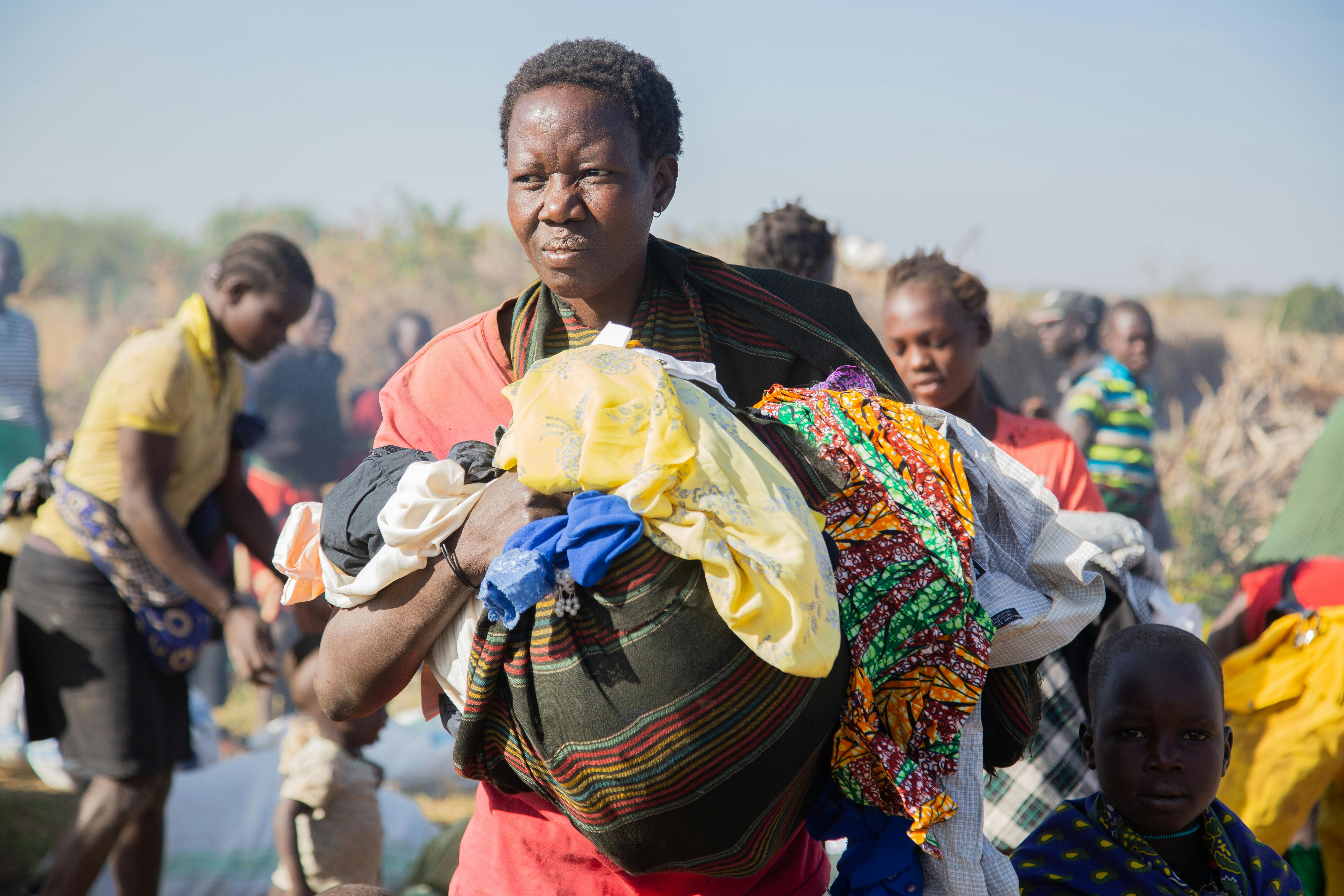 Woman carrying clothing and belongings while shifting through a crowded displacement camp with children nearby