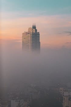 A tall skyscraper rises above dense fog during a colorful sunrise, creating a surreal cityscape.