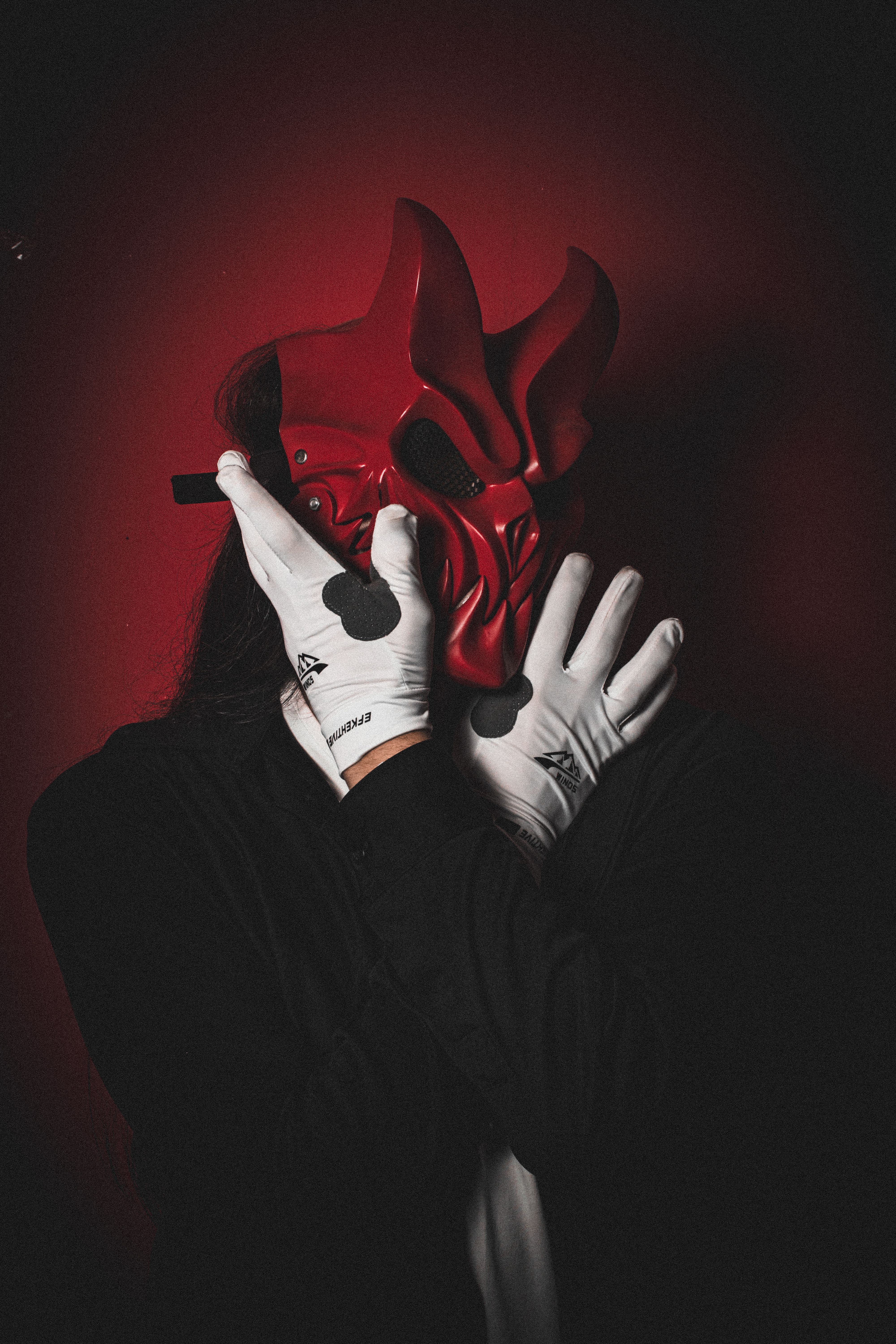 A person wearing a red oni mask and white gloves, set against a dark red background.