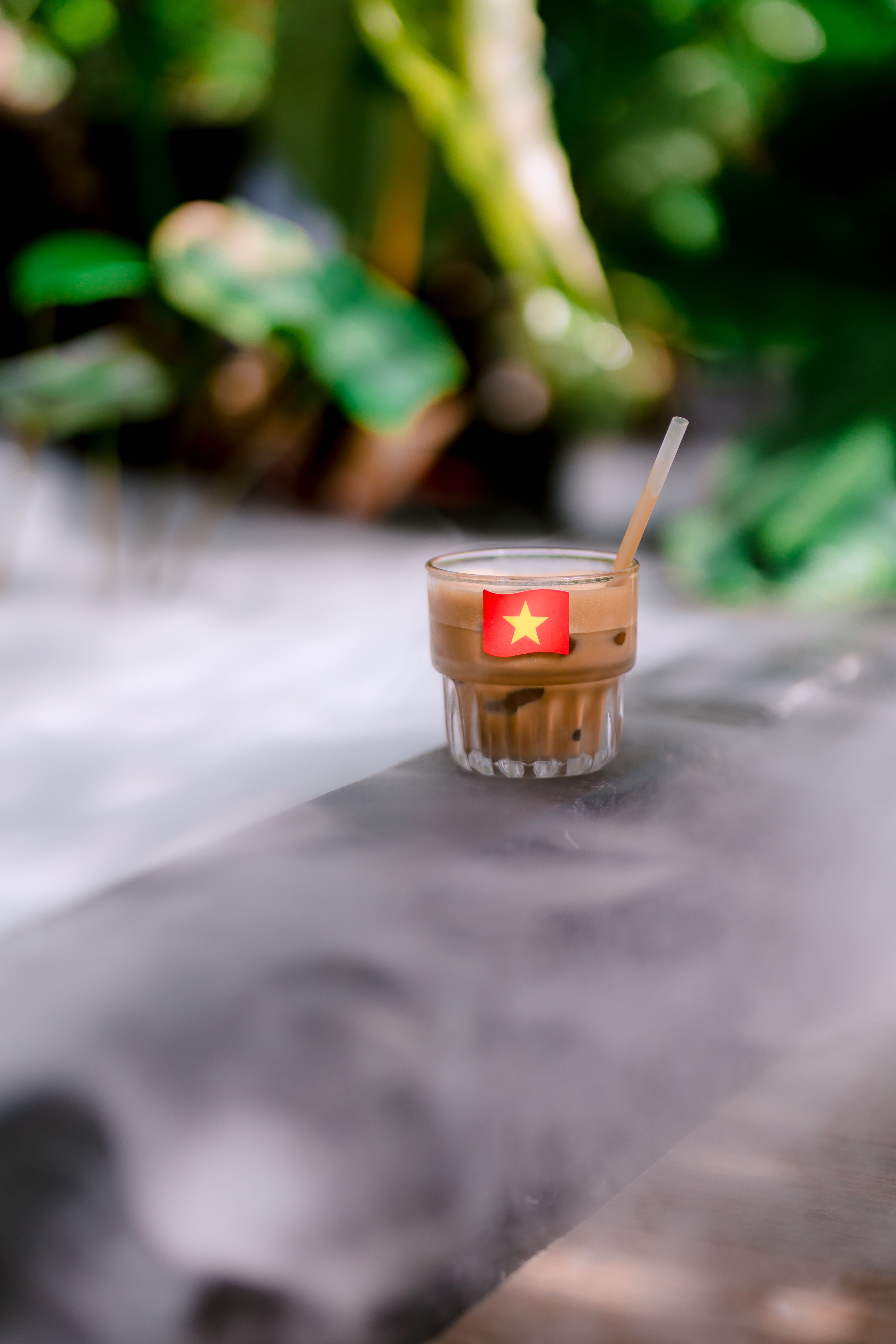 Glass of iced coffee with Vietnamese flag outdoors in leafy surroundings.