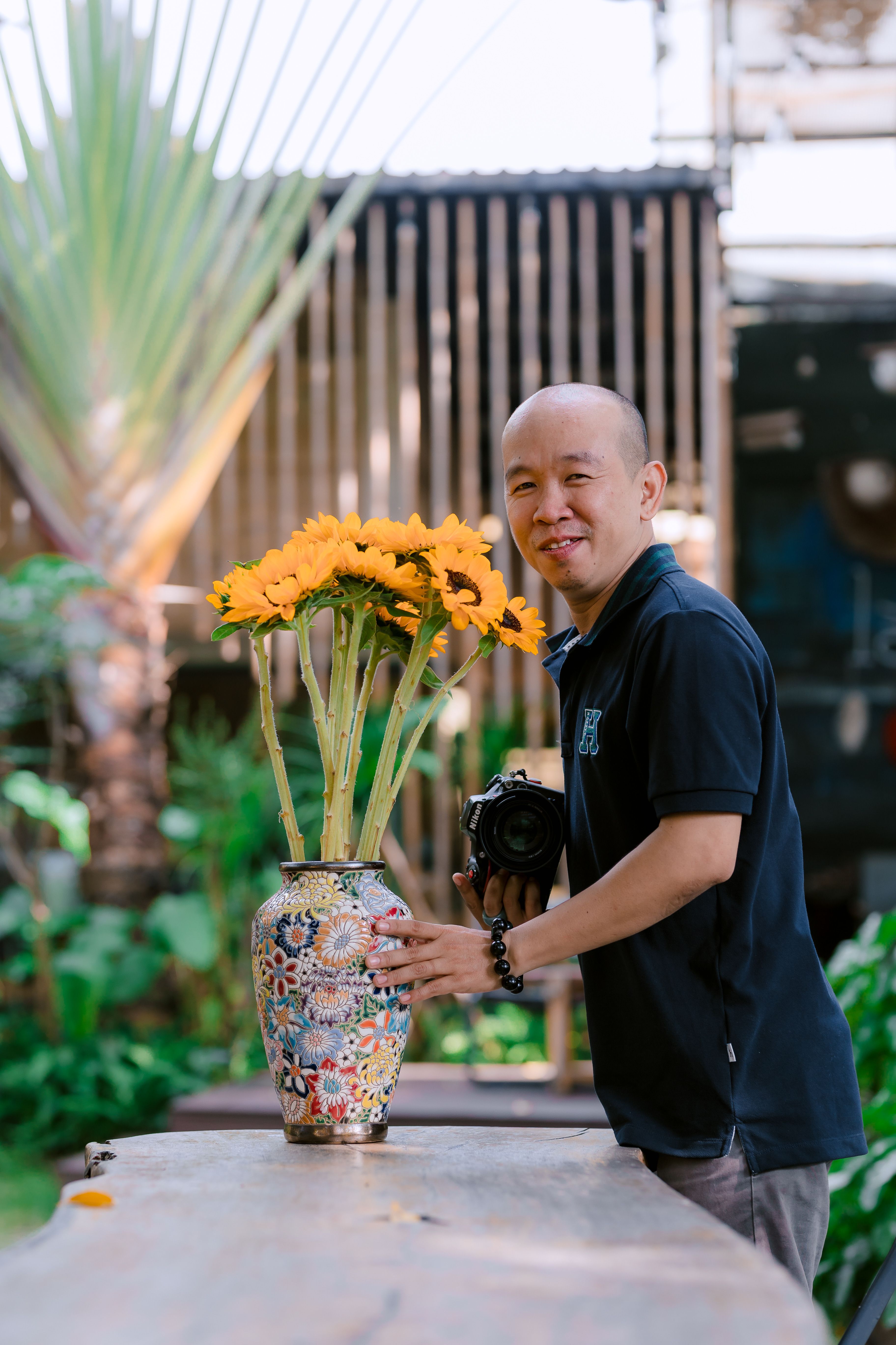 Smiling photographer holds camera beside colorful vase with sunflowers outdoors.