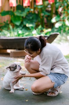 A young woman kneels to interact with a cute French Bulldog in a sunny garden setting.