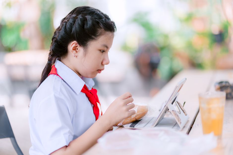学校の女の子が屋外でおやつを食べながらタブレットで勉強し、デジタル学習を具現化しています。