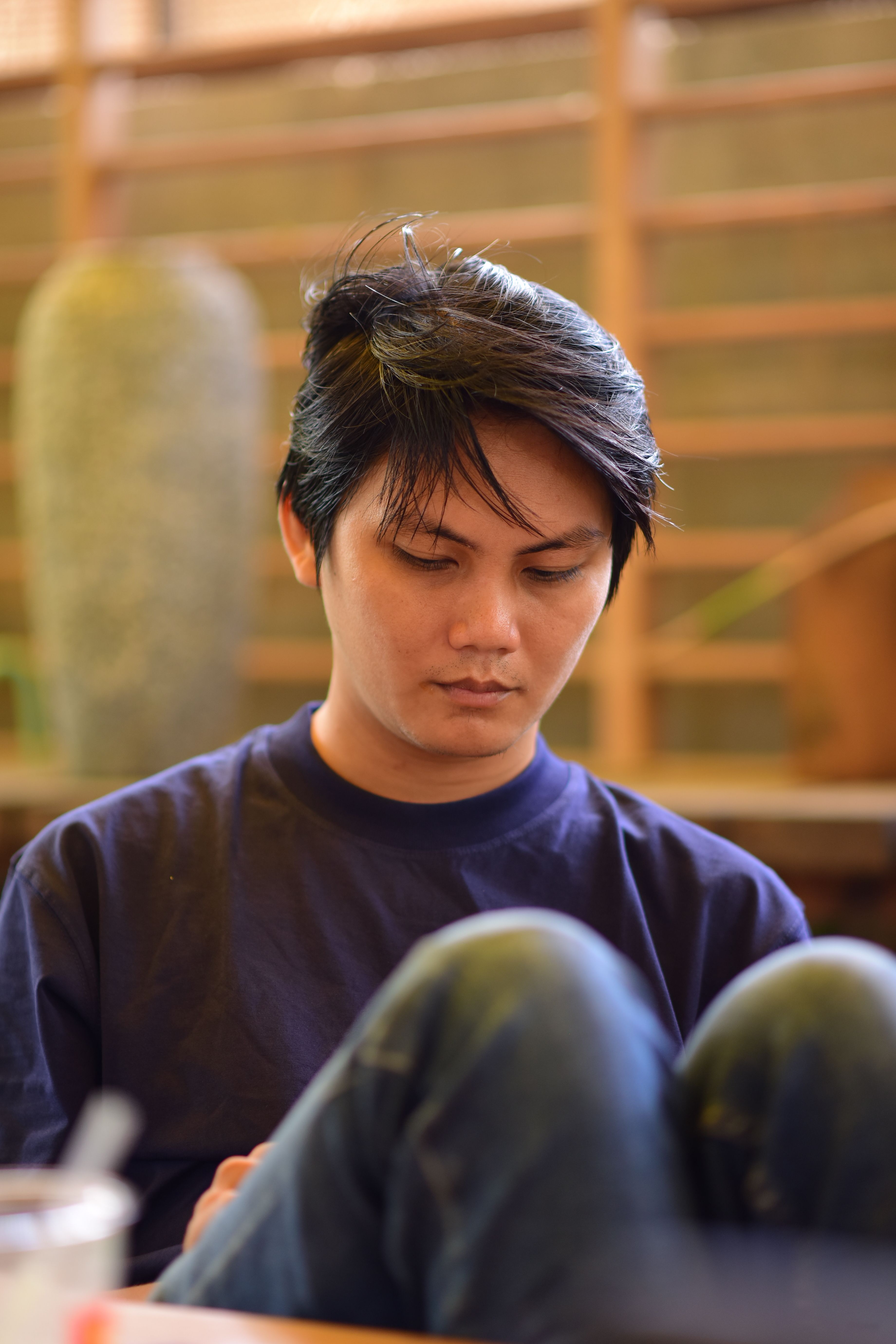 A young man sits indoors, appearing relaxed and thoughtful.