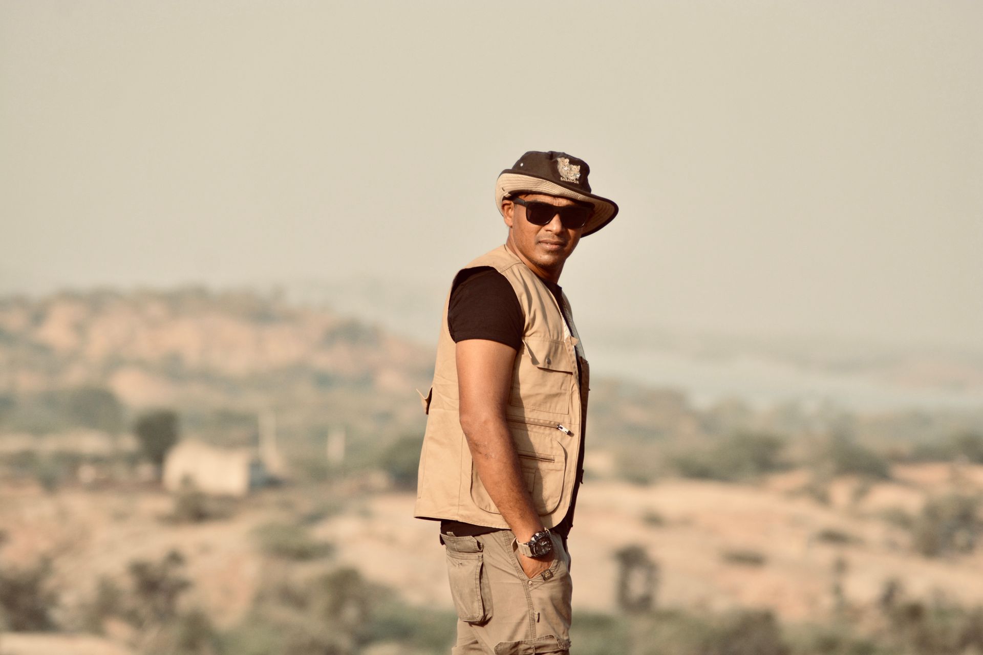 Man wearing safari style attire outdoors in an expansive natural landscape.