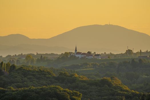 Beautiful landscape of a rural church surrounded by lush greenery at sunrise, with misty mountains in the background.