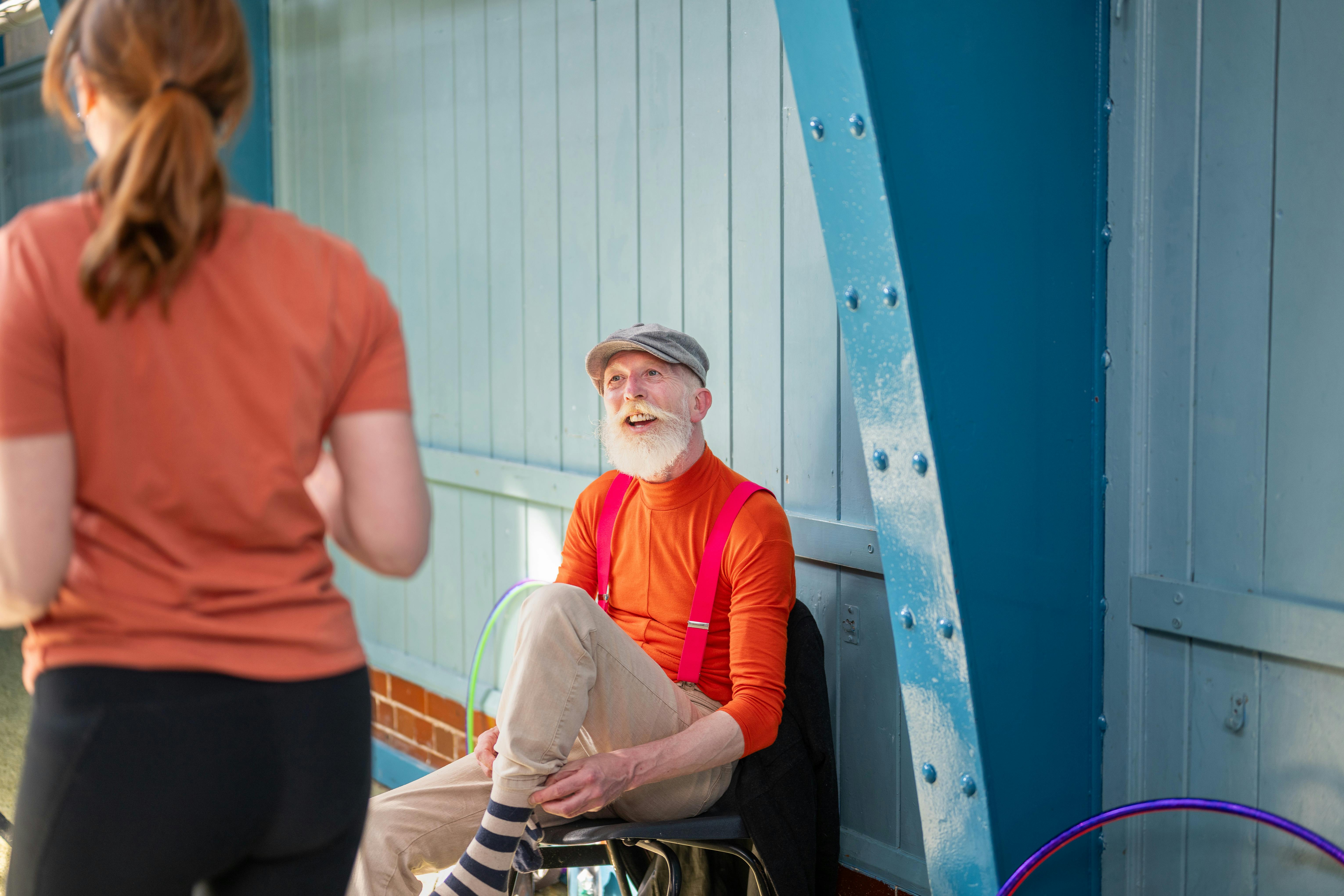 A cheerful elderly man talks with a young woman near a colorful wall, wearing bright orange and suspenders.