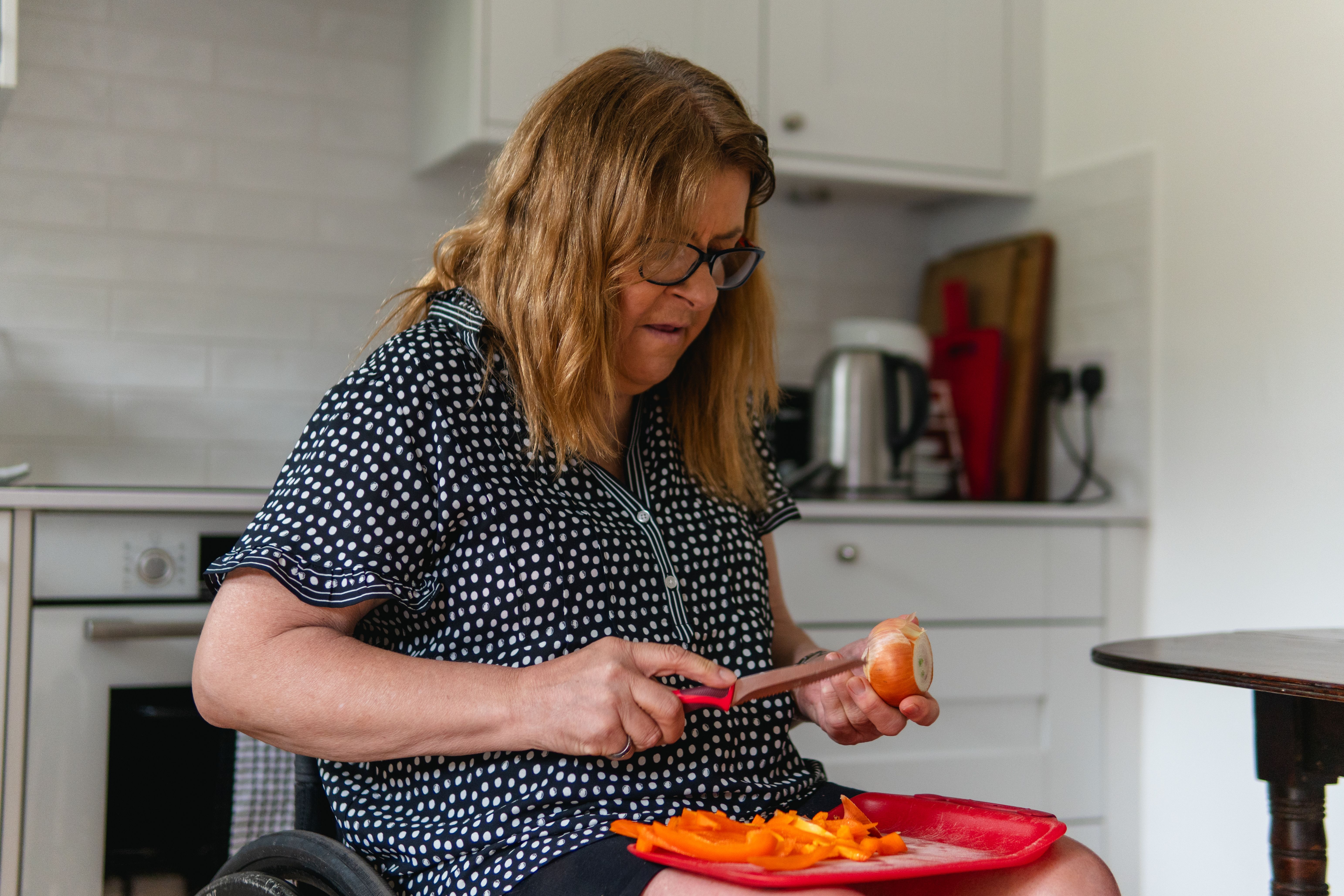Une femme d'âge moyen coupe des légumes dans la cuisine alors qu'elle est assise dans un fauteuil roulant.
