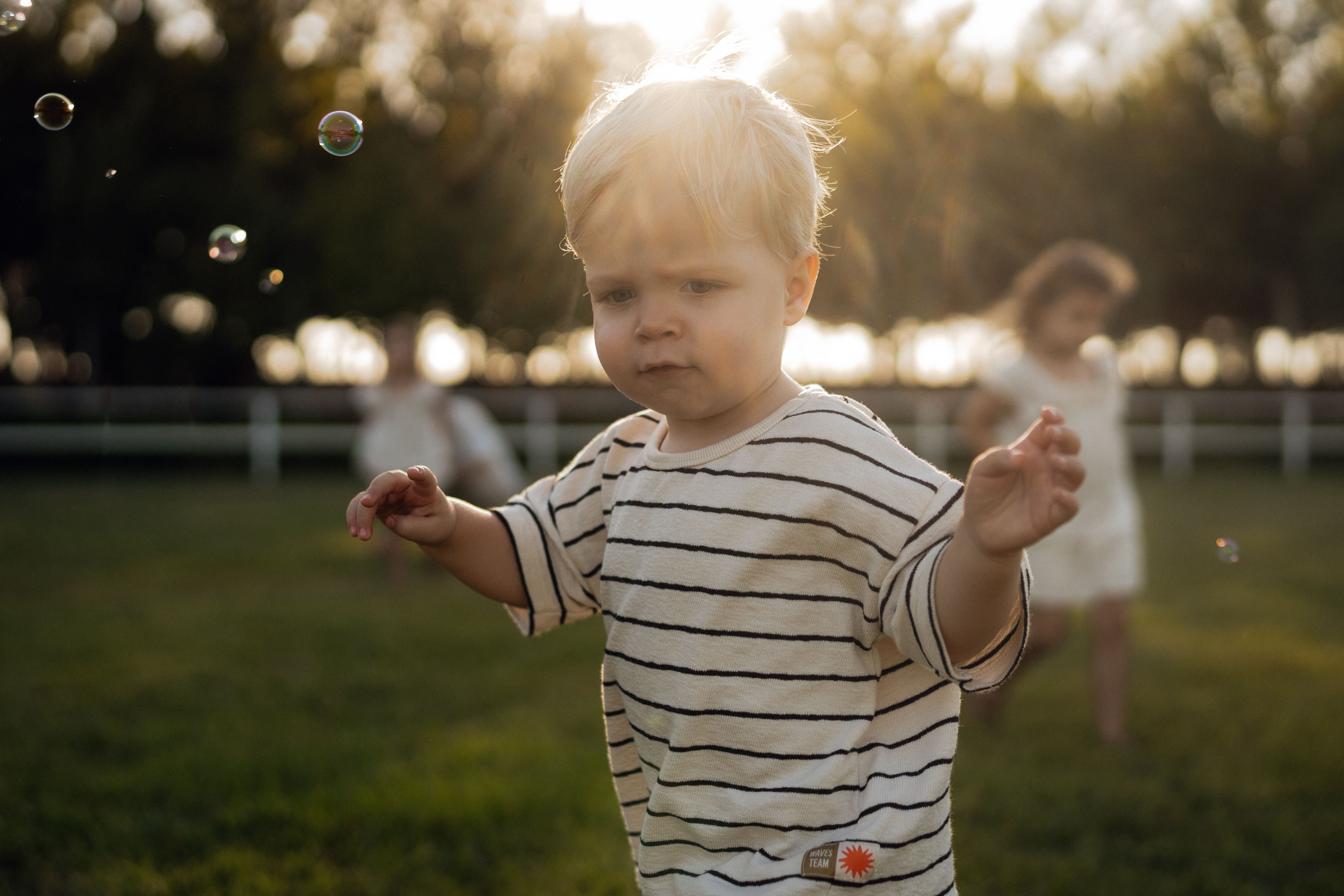A toddler in a striped shirt enjoys playing with bubbles in a sunny park setting.