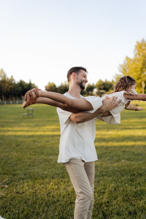 Free A joyful father spins his daughter in a lush green park under a clear summer sky. Stock Photo
