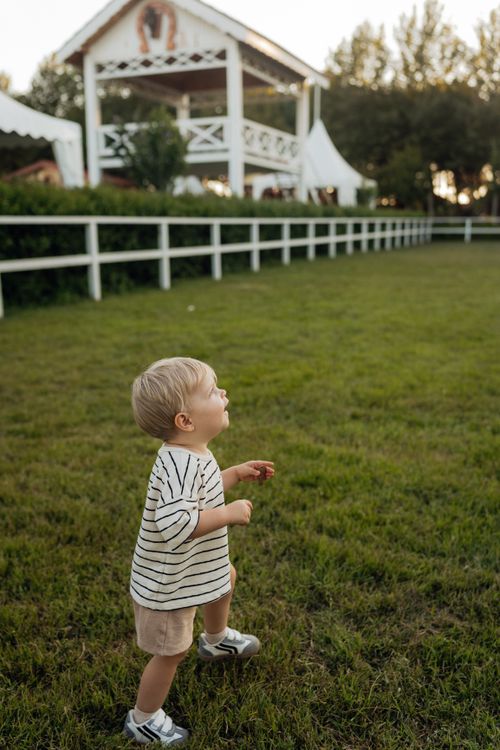 Free Blond toddler in striped shirt joyfully plays on grass near a barn-like building. Stock Photo
