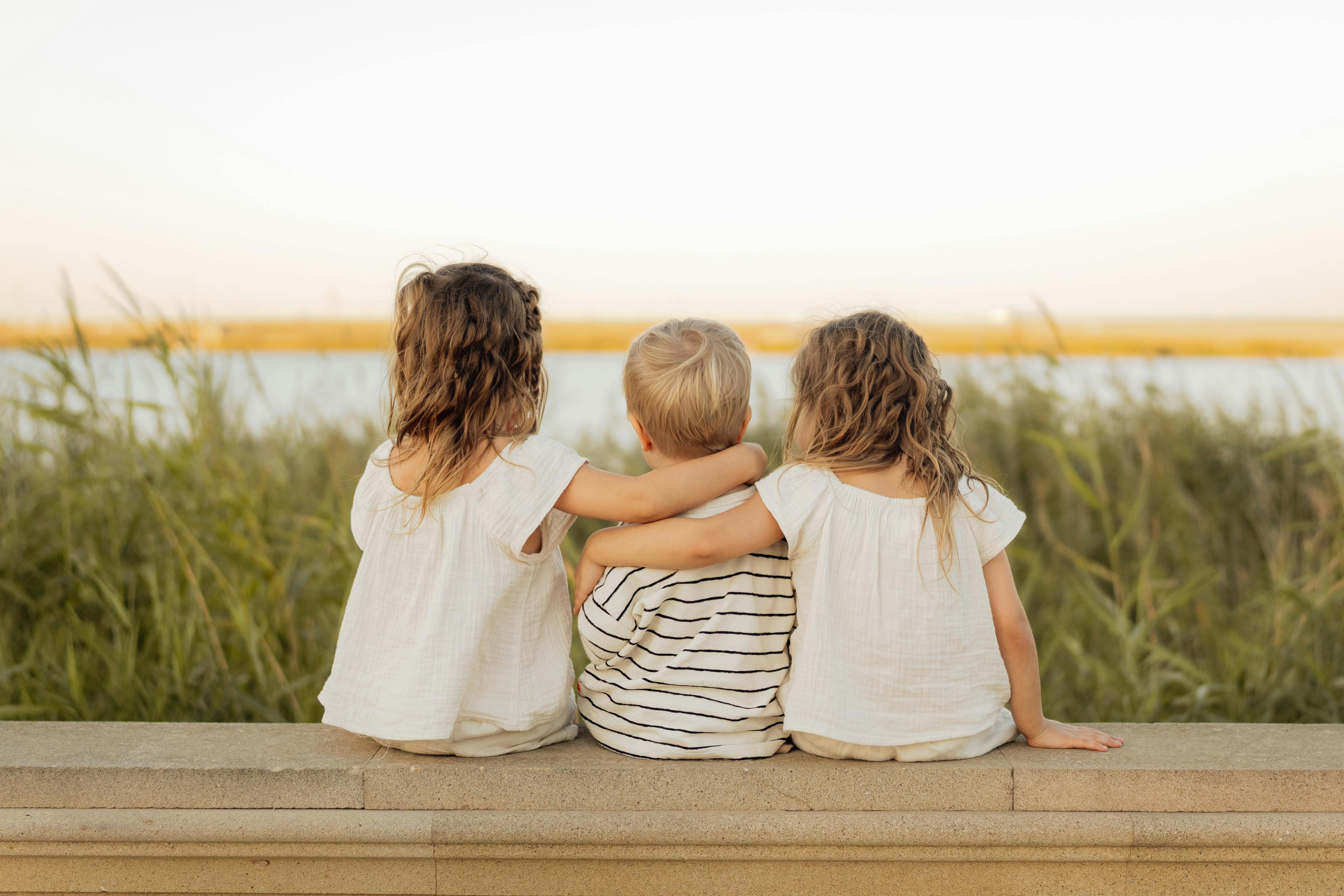 Three children sit on a stone ledge, gazing at a serene lakeside scene, embodying friendship and tranquility.