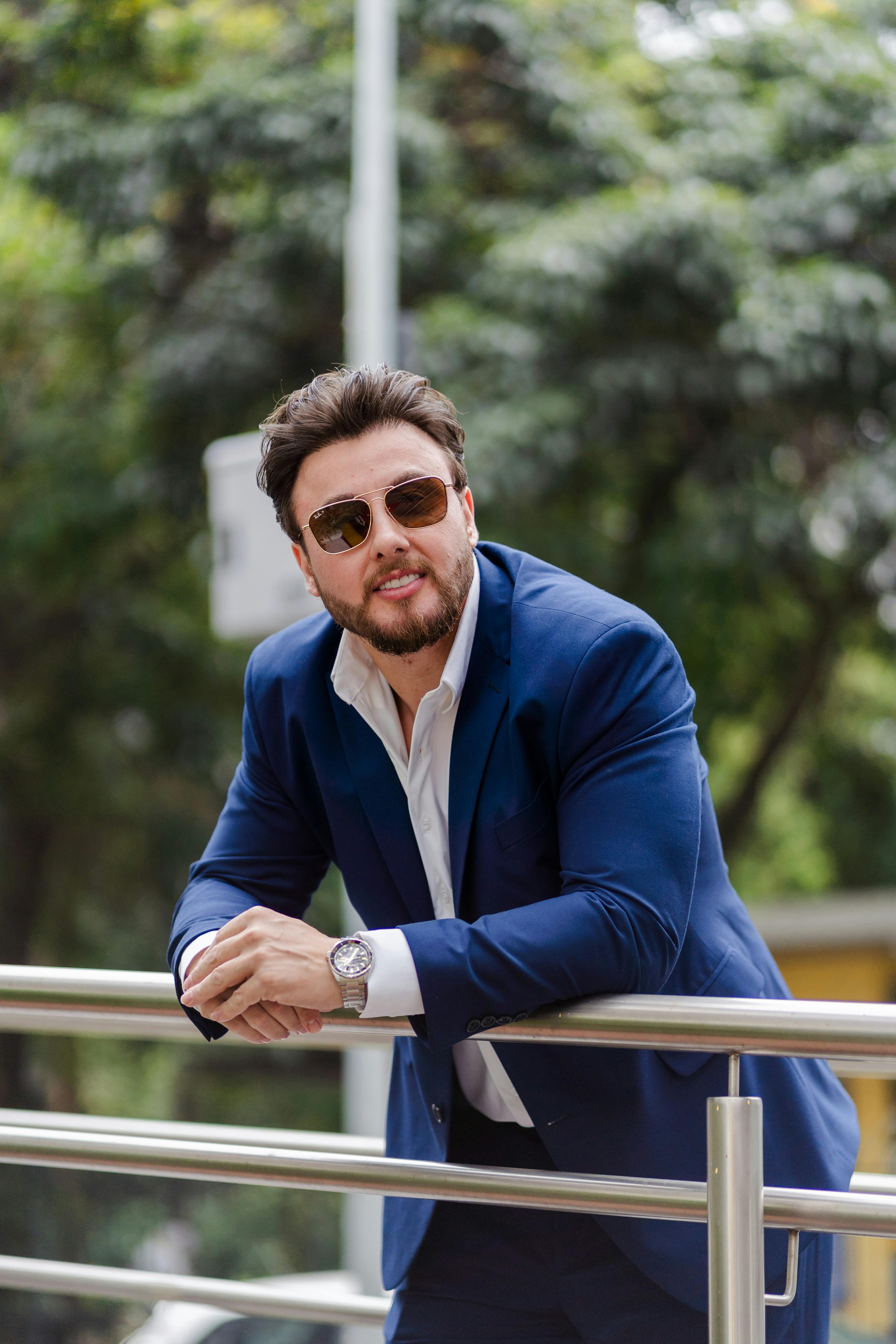 Smiling man in blue suit and sunglasses leaning on railing in an outdoor setting.