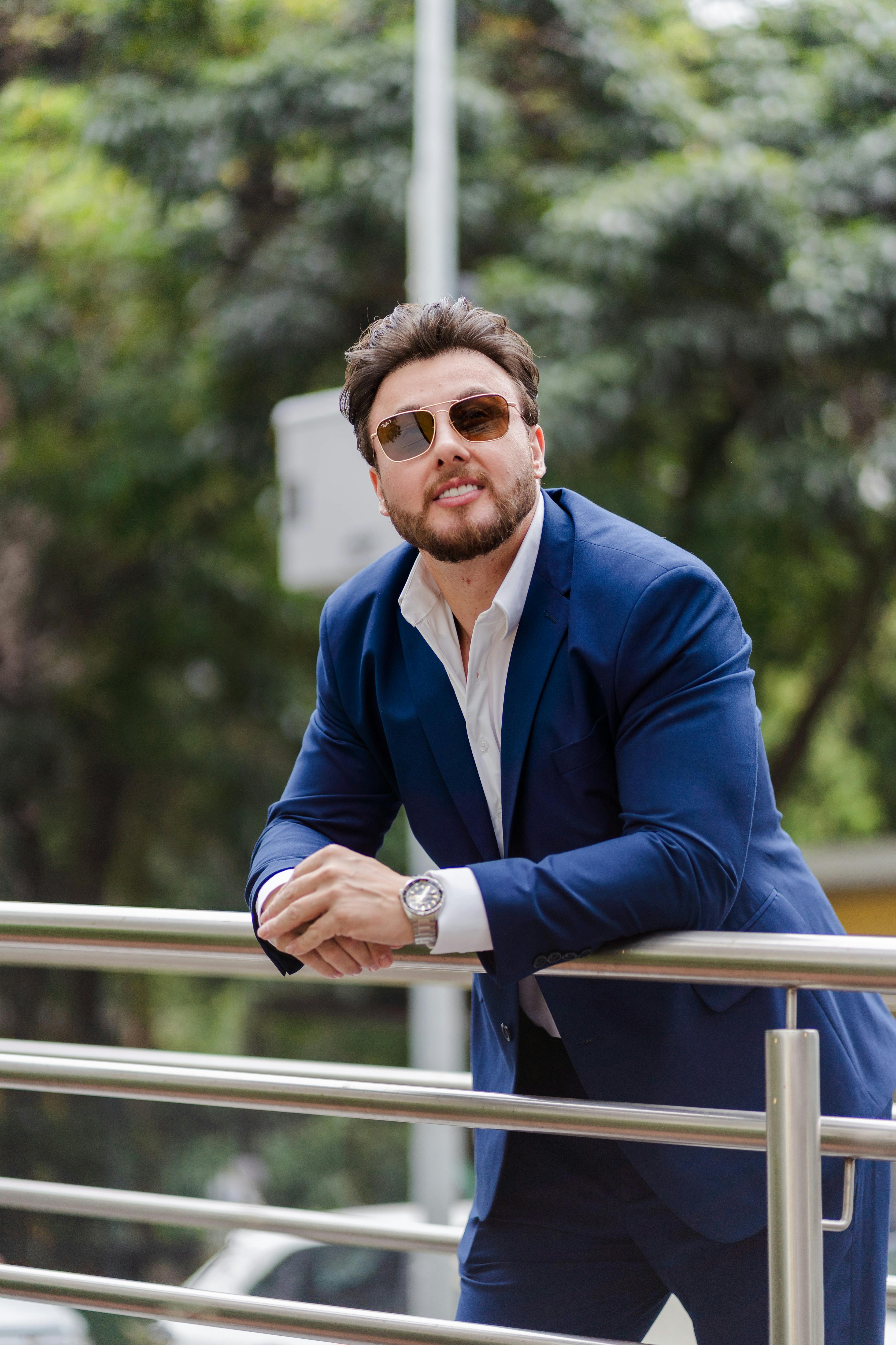 Stylish man in sunglasses leaning on railing, wearing a blue suit outdoors.