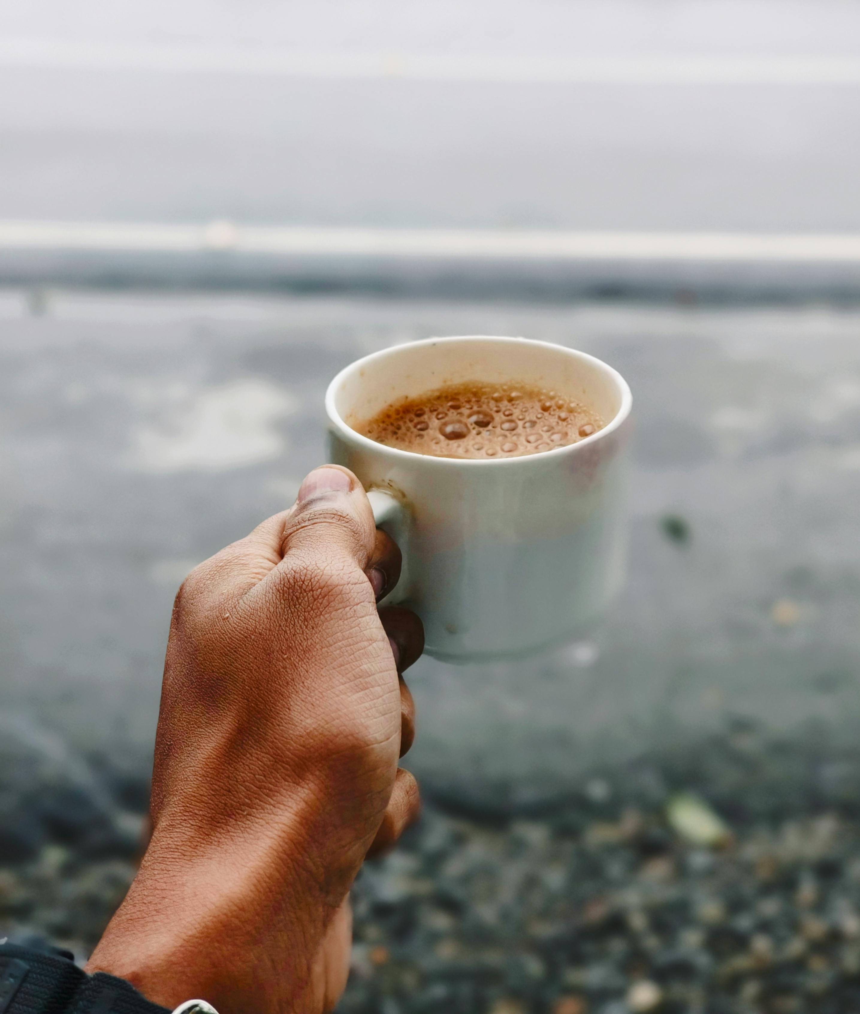 A hand holding a steaming cup of tea outdoors, captured on a cloudy day in India.