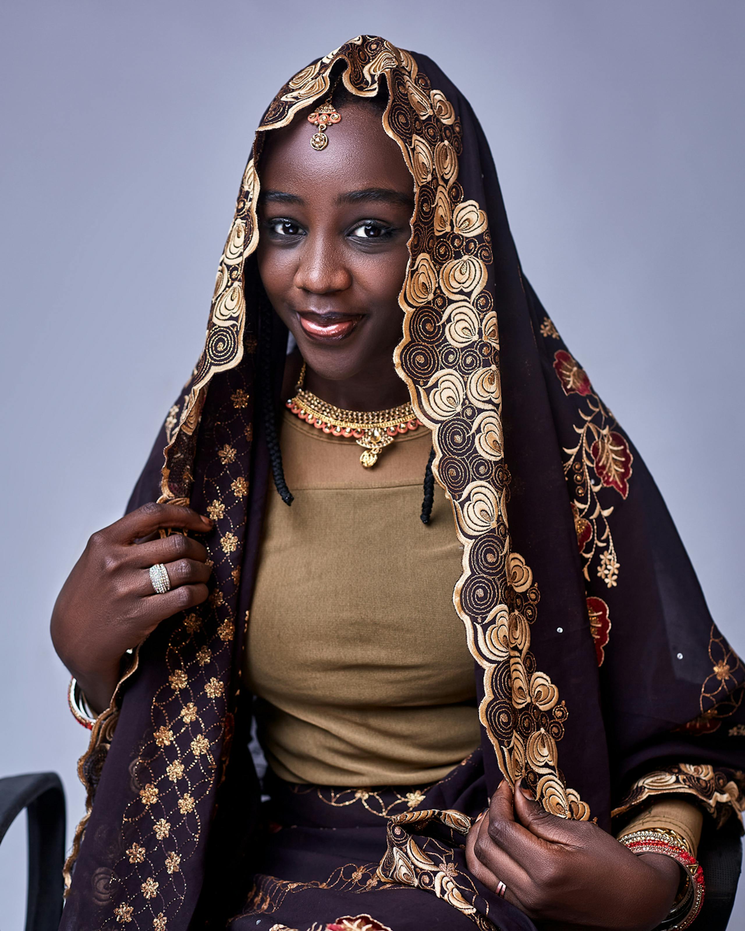 Portrait of a Nigerian woman in traditional attire wearing a beautifully embroidered headscarf, smiling warmly.