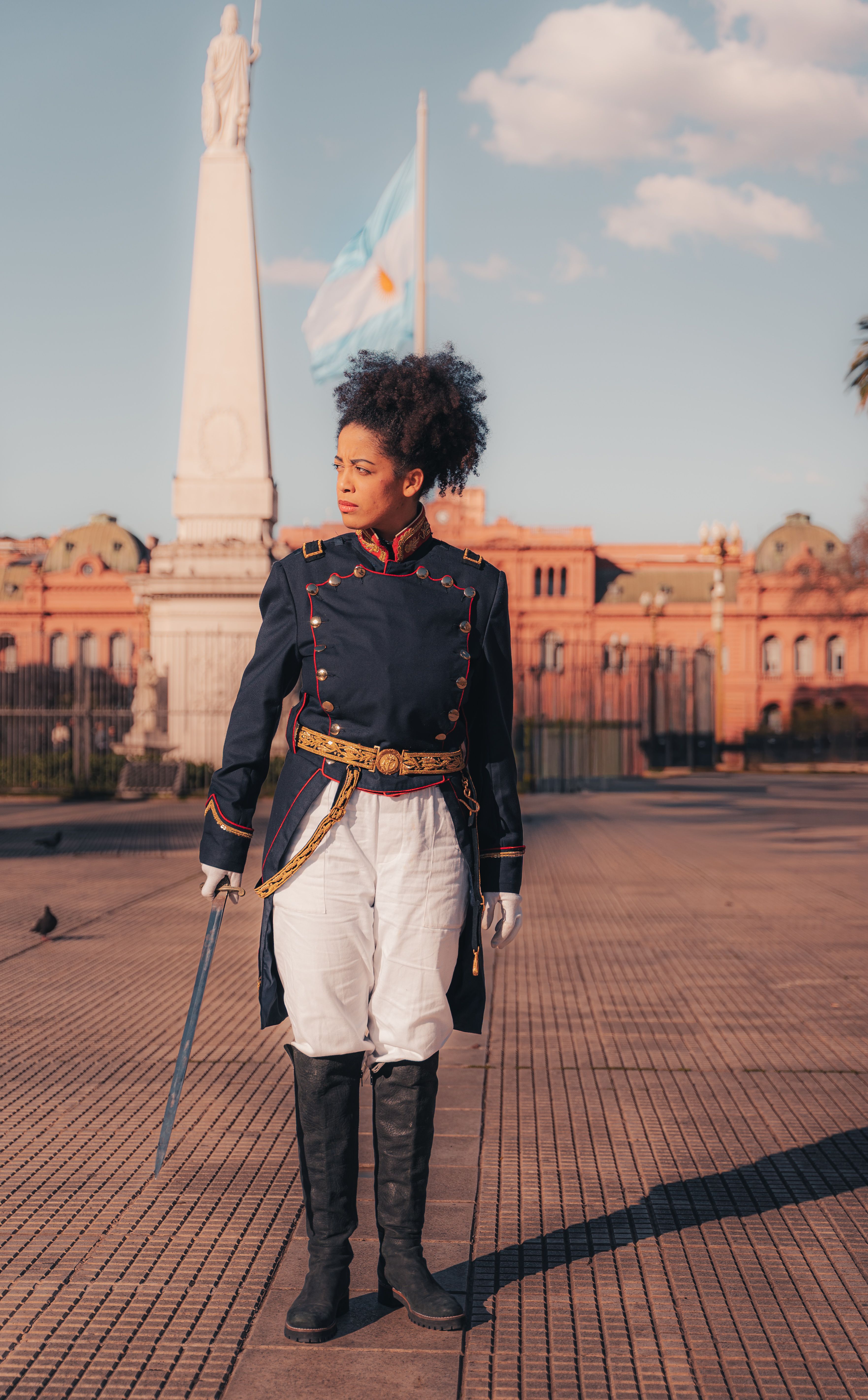A person in traditional military uniform stands with a sword in front of a monument on a sunny day.