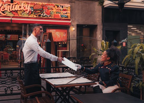 A waiter serves a menu to a seated customer at an outdoor restaurant with colorful signage.