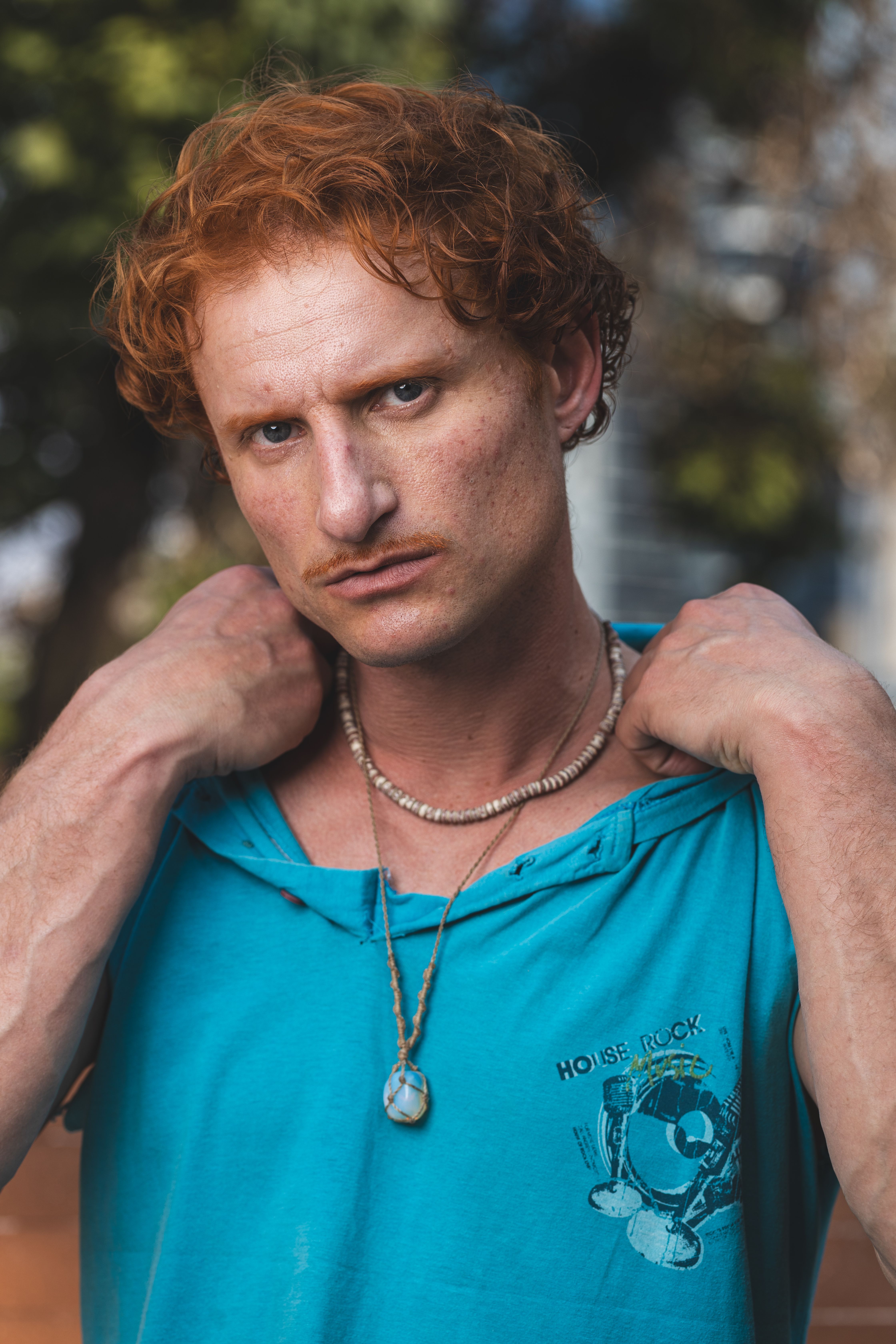 Portrait of a confident man with curly red hair in a blue shirt outdoors, wearing jewelry.
