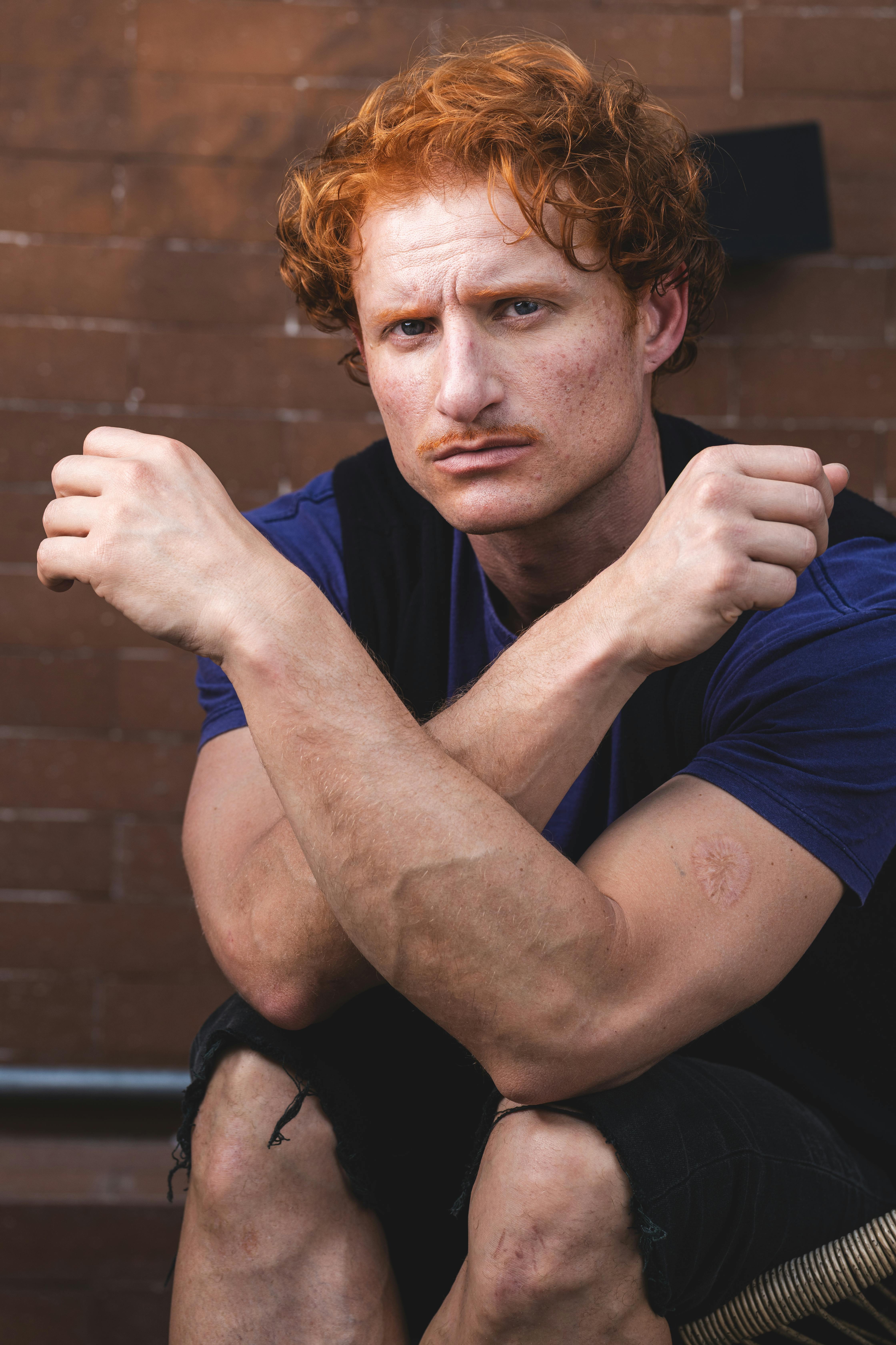 Red-haired man with mustache sitting outdoors, arms crossed, looking confident. Casual attire against a brick background.