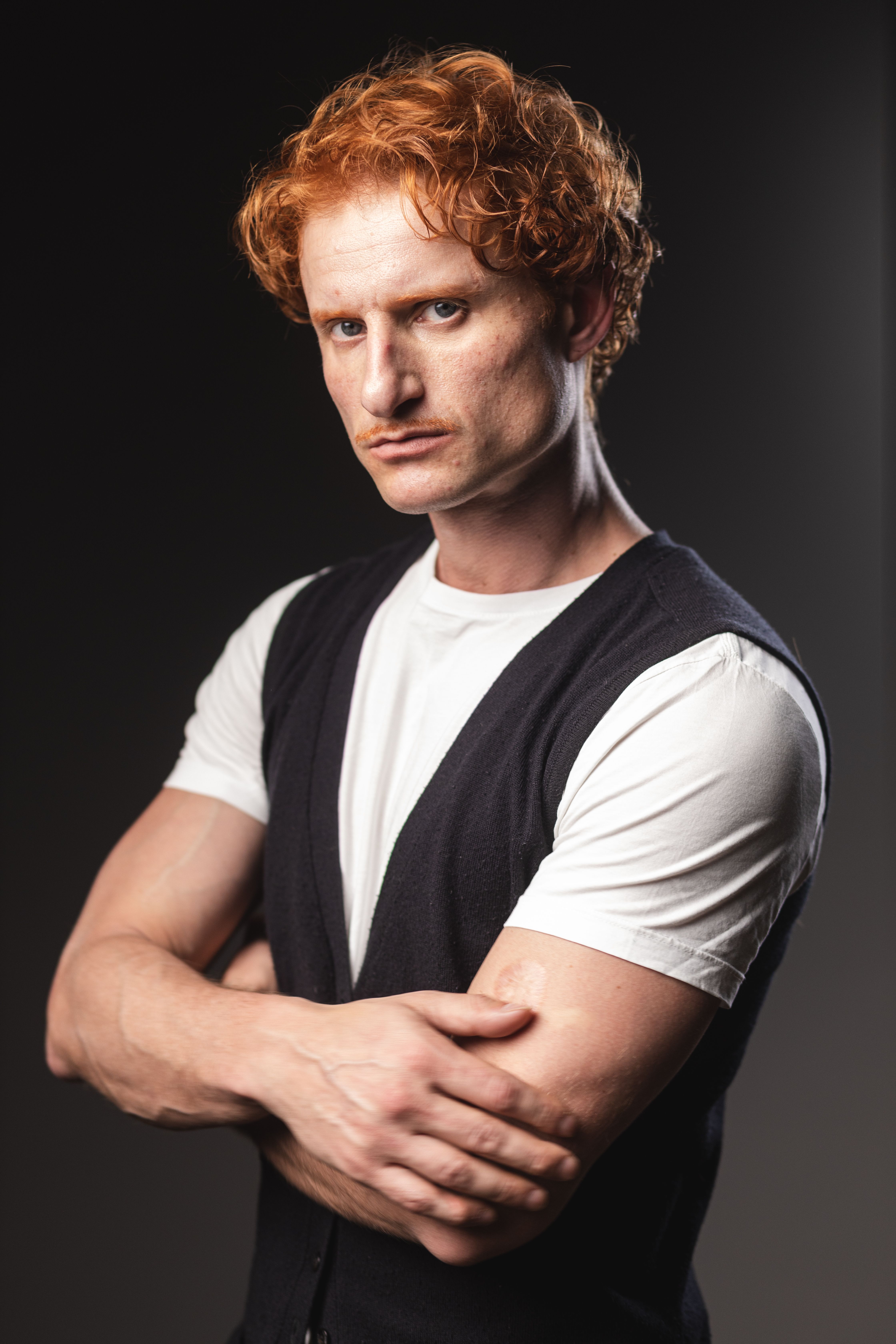 Stylish young redhead man posing confidently in a studio setting, wearing a casual vest and t-shirt.