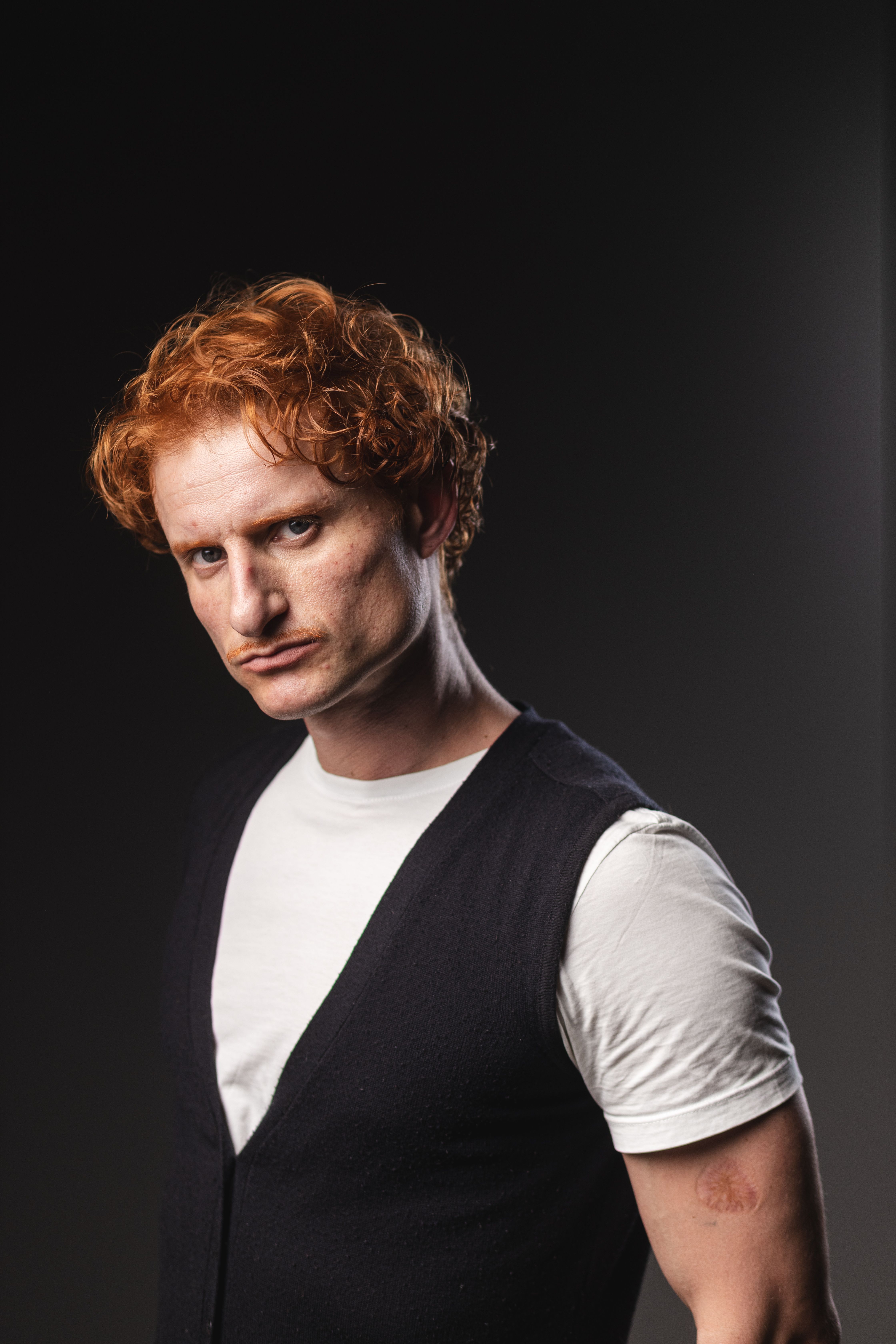 Close-up portrait of a redheaded man in a vest and white shirt, dramatic lighting.