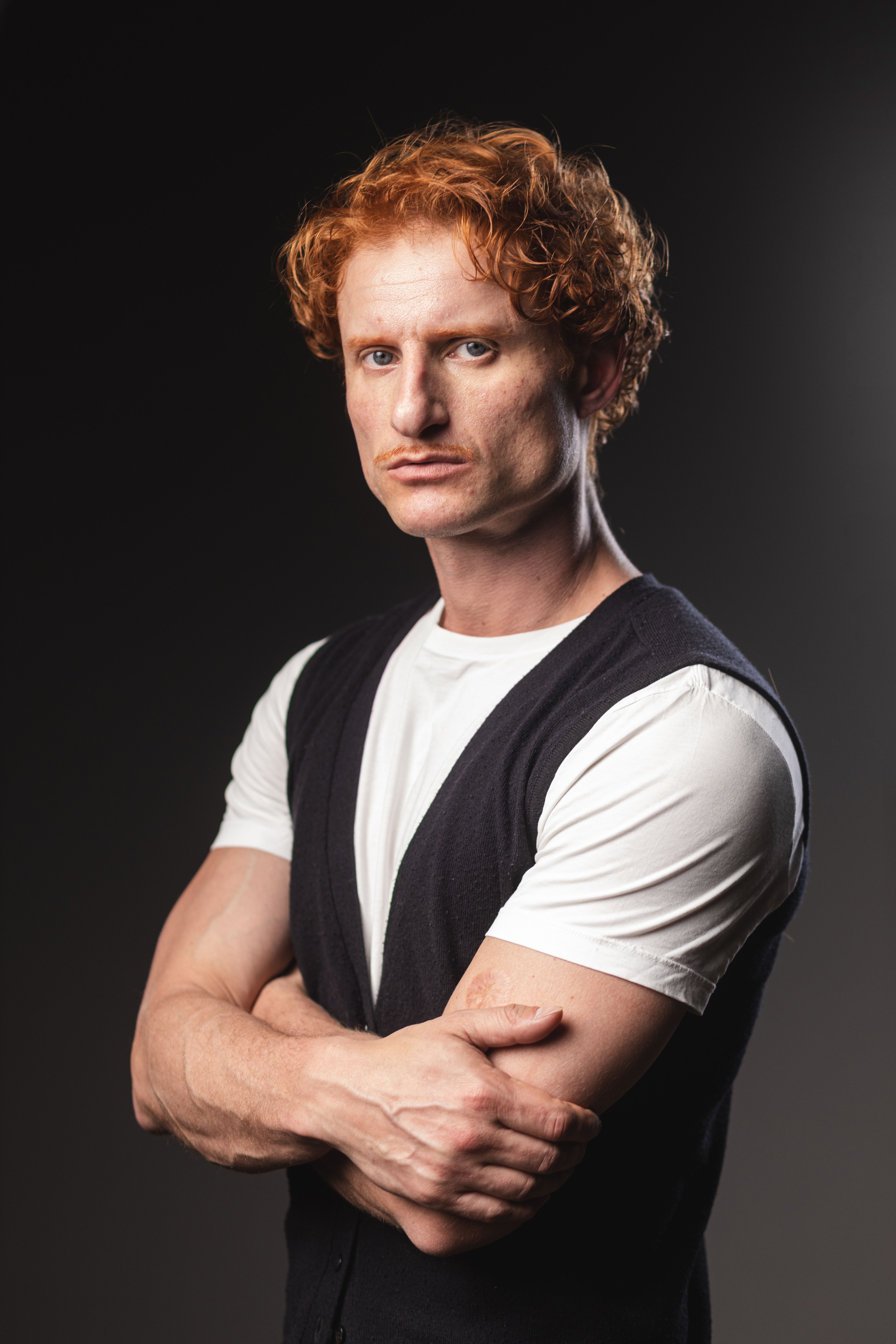 Confident red-haired man poses with crossed arms in a studio shot.
