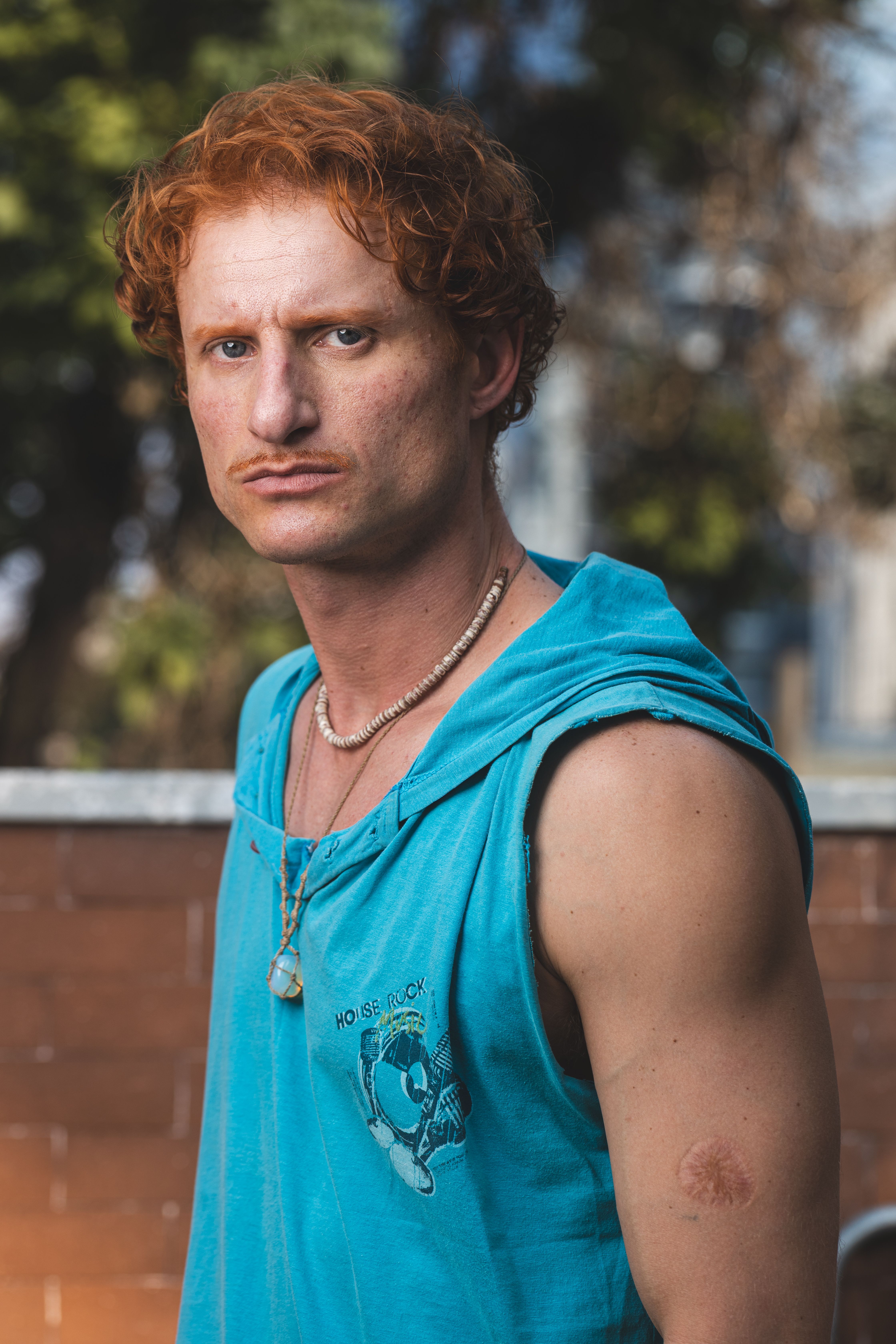 A red-haired man in a blue sleeveless hoodie with a necklace poses outdoors, looking directly at the camera.