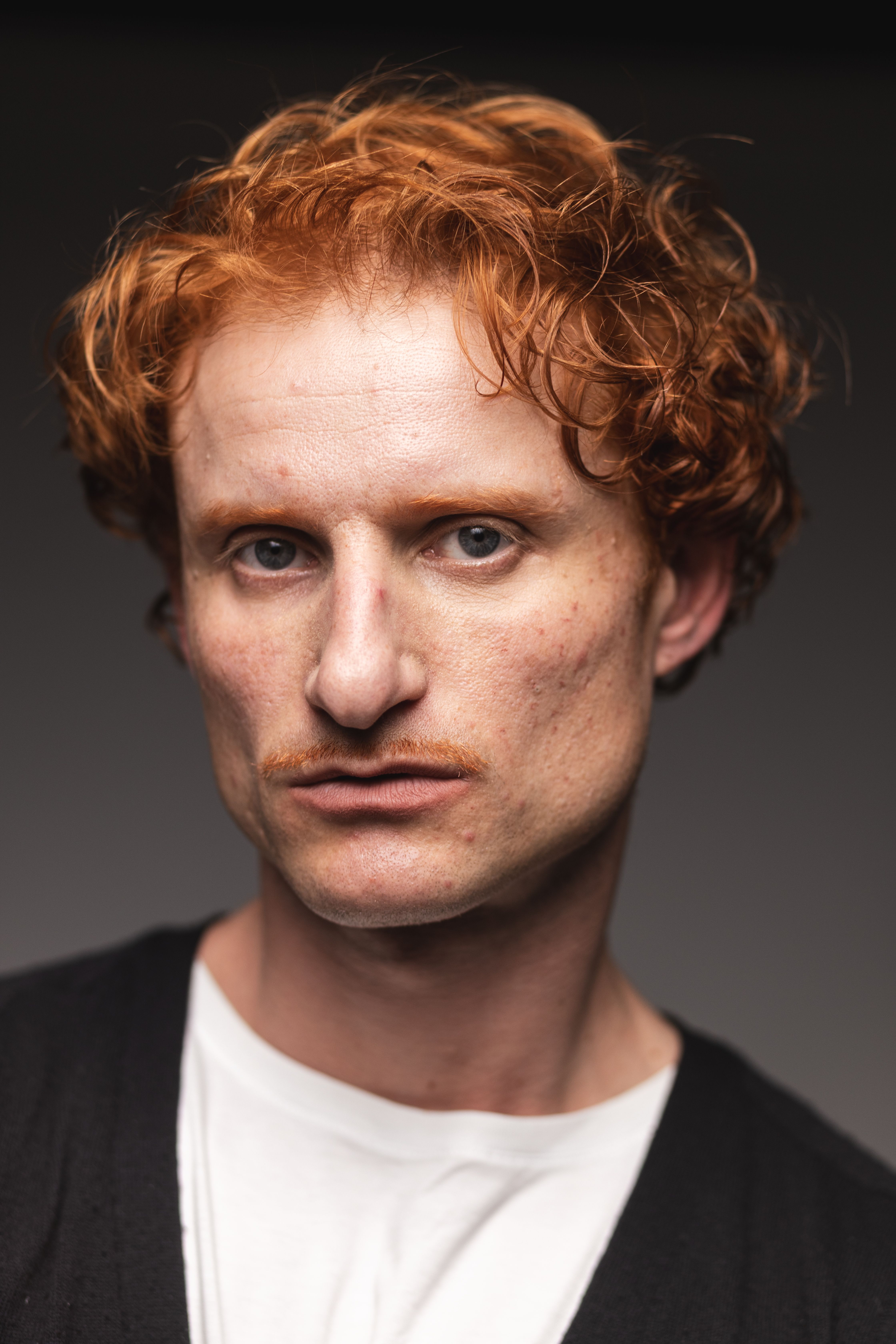 Close-up portrait of a man with curly red hair and blue eyes against a neutral background.