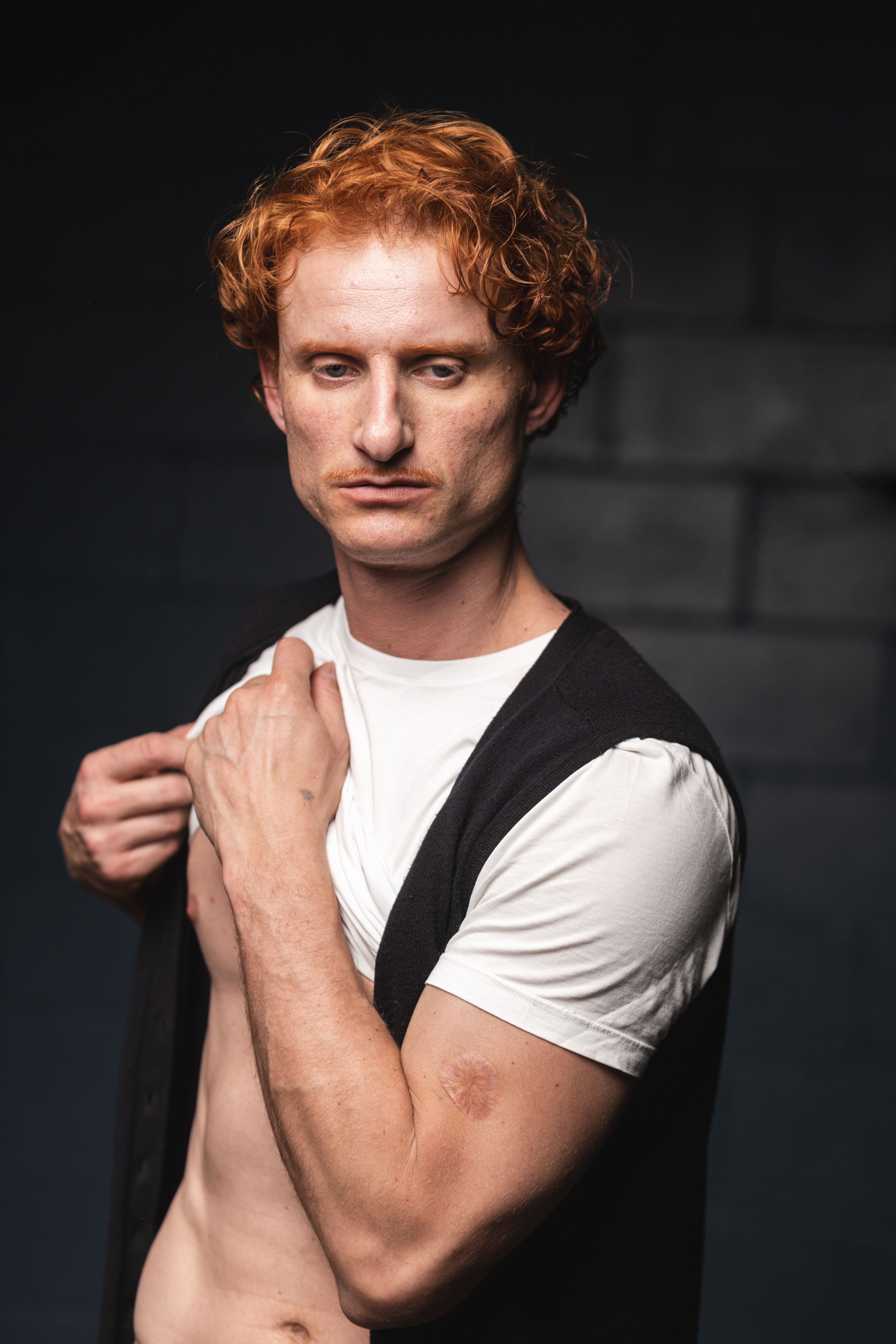 Redheaded man wearing a white shirt and black vest posing indoors against a dark background.