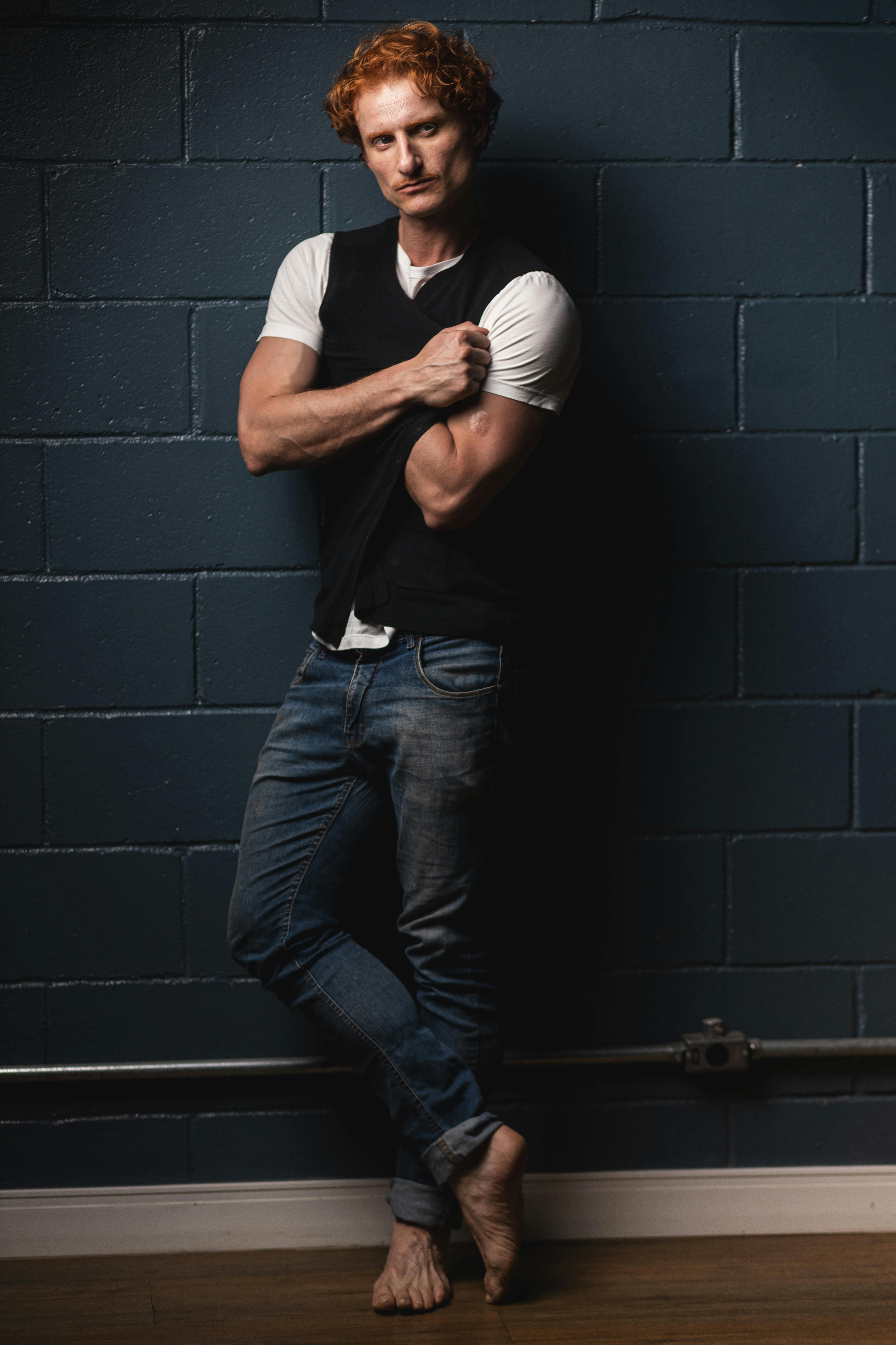 Confident man with red hair poses barefoot against a dark indoor wall, exuding modern relaxed style.